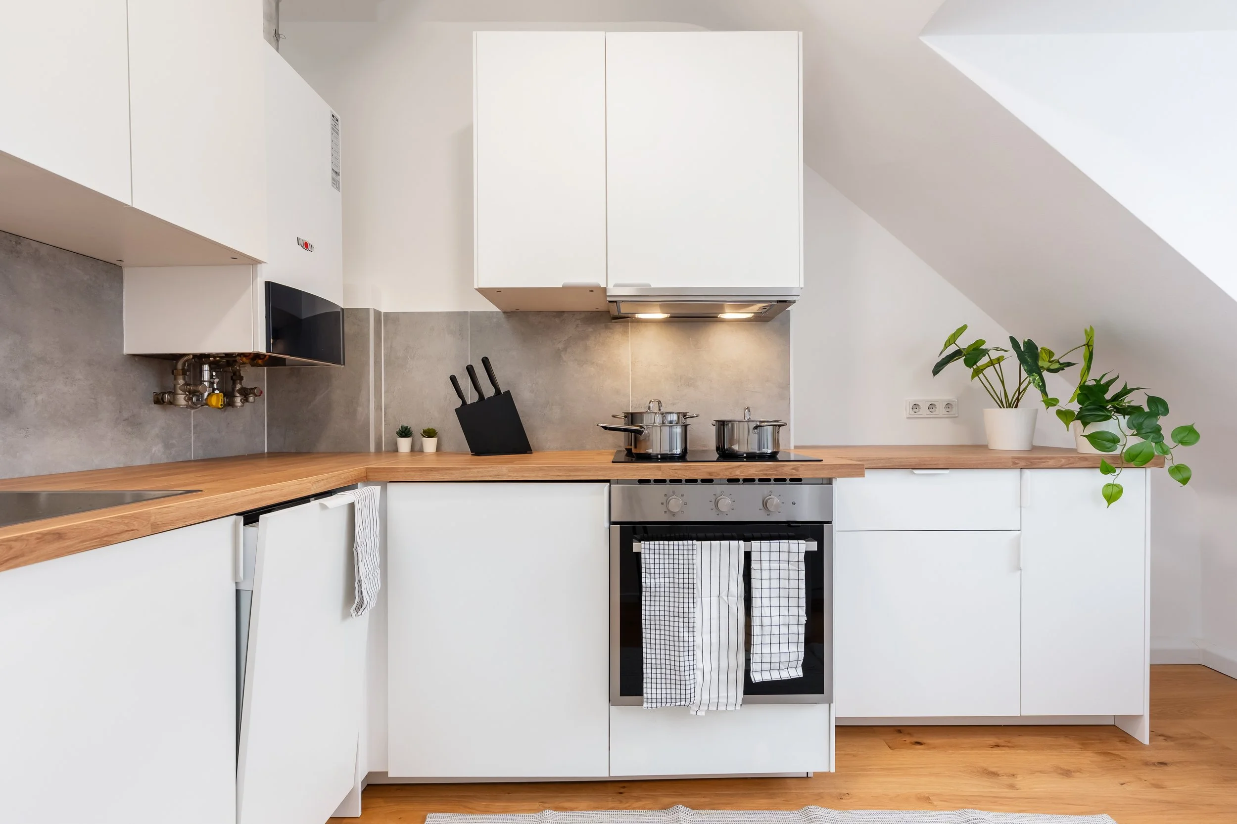 Moderne Küche mit weißen Schränken, Holzarbeitsplatte, Herd mit Topf und Warnschild, Küchenutensilien, zwei kleine Pflanzen, eine größere Pflanze in einer weißen Tüte, graue Wandfliesen und Holzboden.