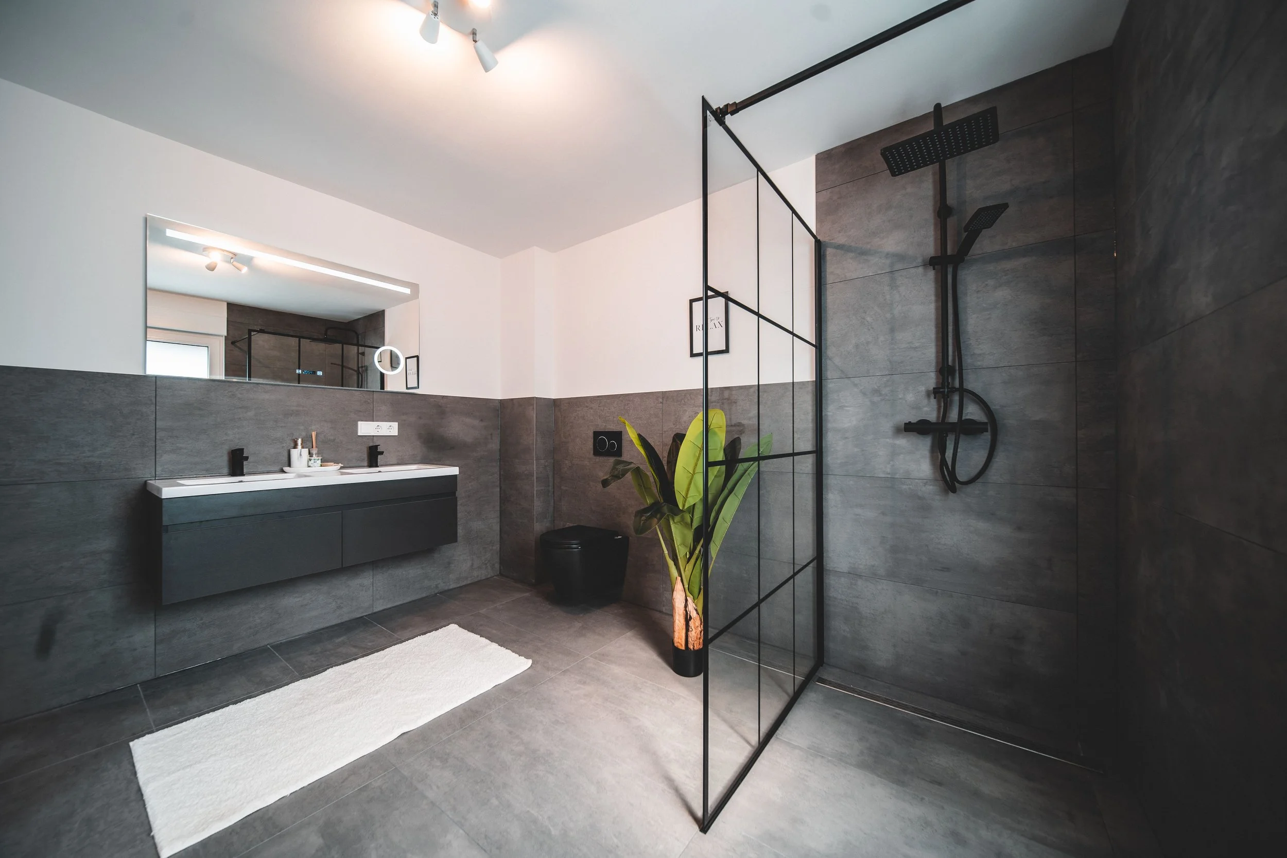 Moderner Badezimmer mit dunklem Waschtischuntrag, großem Spiegel, Handtuch, Pflanze und begehbarer Dusche mit schwarzen Armaturen.