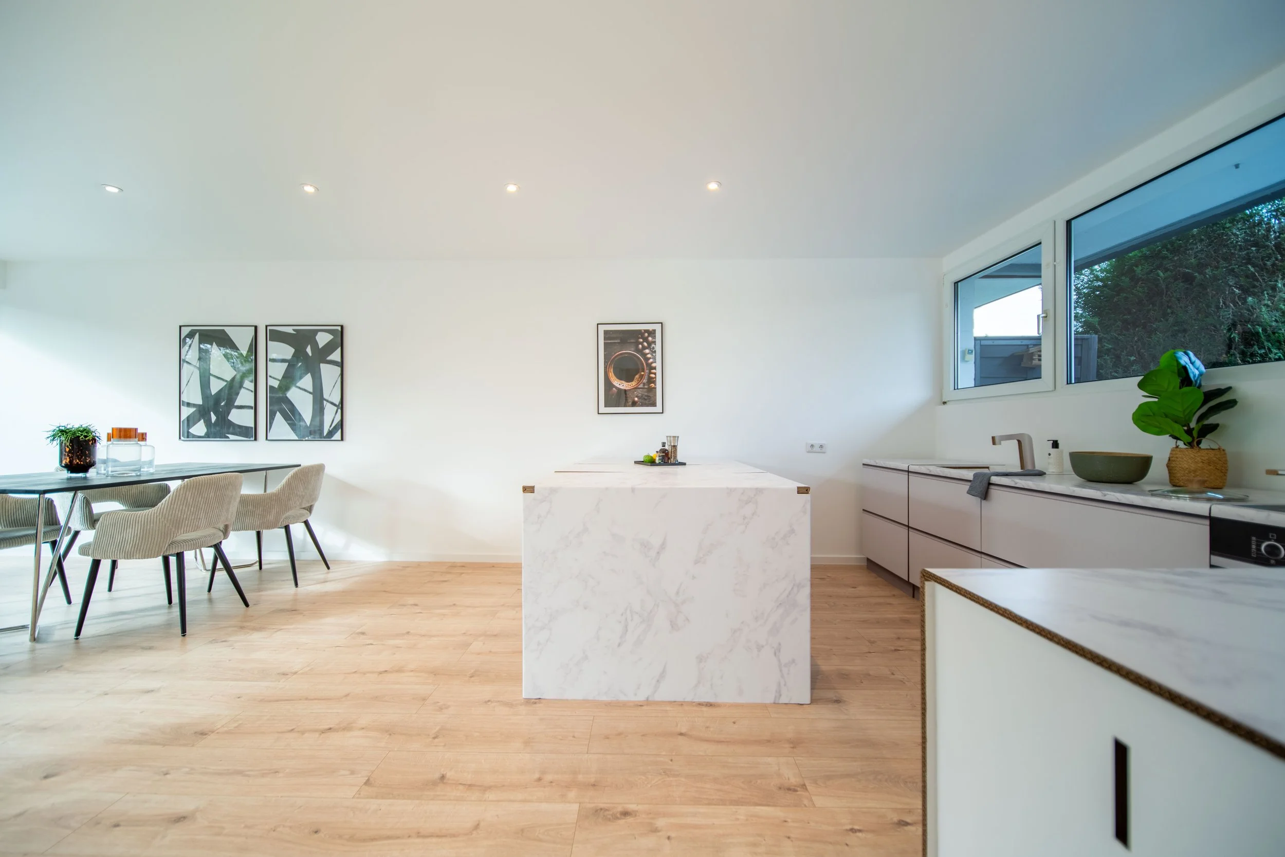 Moderne, helle Küche mit weißen Schränken, großen Fenster und Marmorinsel, Holzfußboden, Wandkunst und Pflanzen.