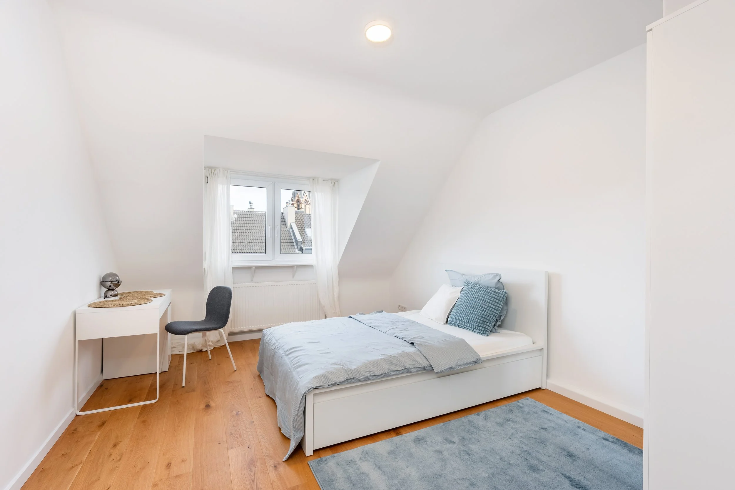 Schlafzimmer mit Weißem Bett, grauen Kissen, blauer Decke, Holzboden, Fenster mit weißen Vorhängen, weißen Wänden, Teppich auf dem Boden, kleiner Schreibtisch, grauer Stuhl, Deckenleuchte