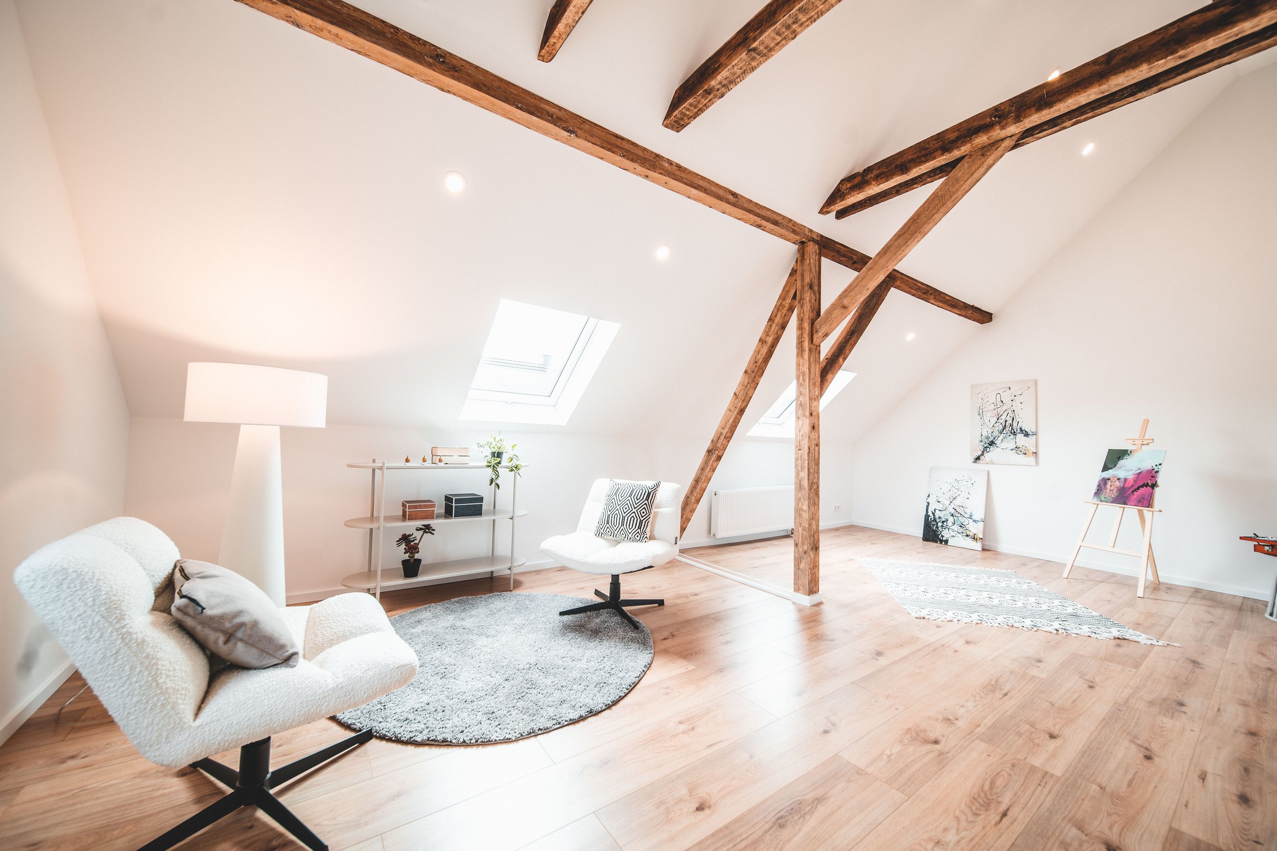Ein modernes, helles Dachzimmer mit Holzfußboden und sichtbareren Holzbalken an der Decke. Es gibt einen weißen Sessel, einen weißen Stuhl, eine Stehlampe, eine kleine Regalwand mit Pflanzen und Dekorationen, sowie einige Gemälde und eine Staffelei a