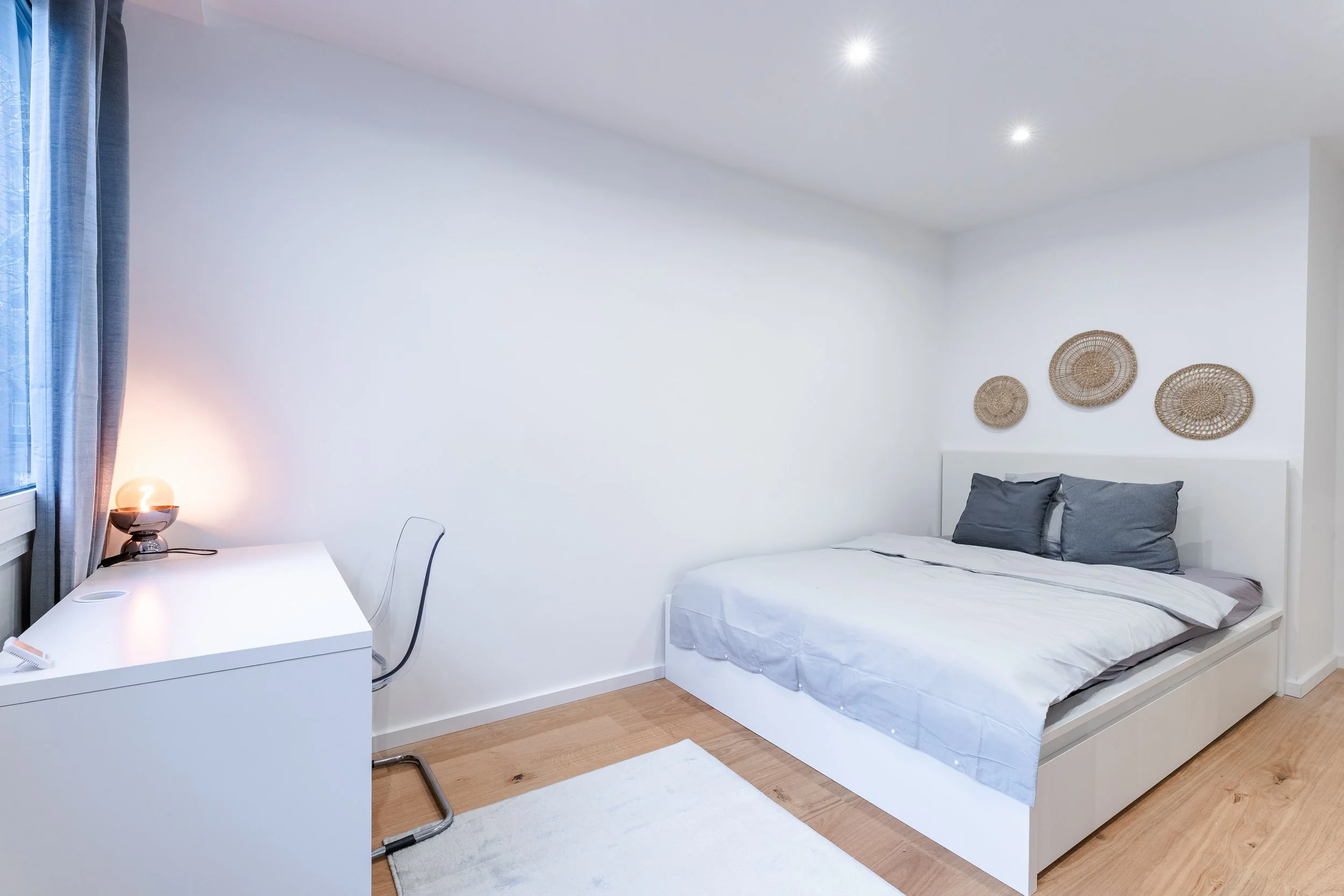 Schlichtes Schlafzimmer mit weißem Bett, dunklen Kissen, Wanddeko aus geflochtenen Körben, Schreibtisch mit Lampe und Stuhl, Holzboden, grauem Vorhang, natürlichem Licht und minimalistischem Design. Kapitalanlagen in Köln, Düsseldorf und Bonn