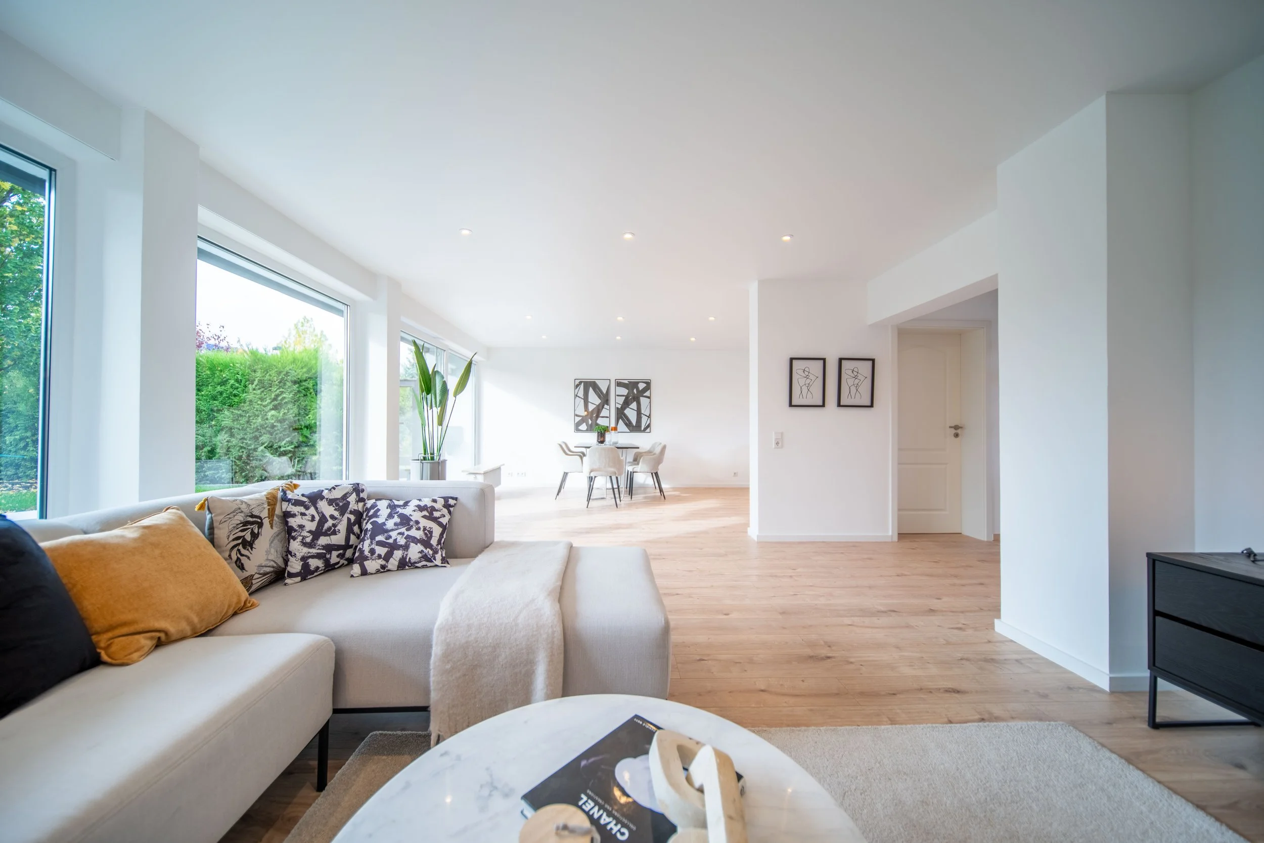 Geräumiges, modernes Wohnzimmer mit großen Fenstern, weißen Wänden, Holzfußboden, Sofas mit Kissen, Couchtisch, abstrakte Wandkunst und einem angrenzenden Essbereich.