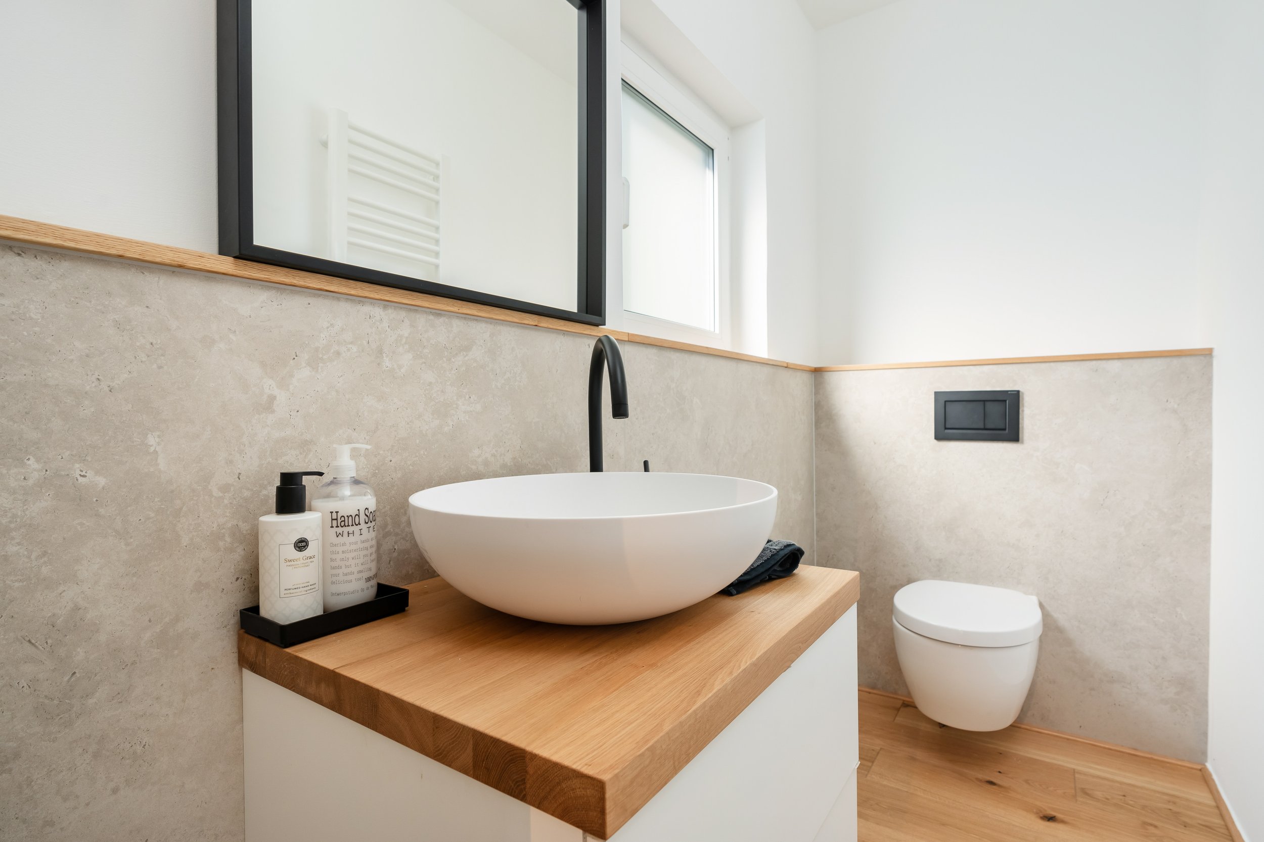 Moderner Badezimmerbereich mit Waschtisch, schwarzem Handwaschmittelspender, Seifenspender und Wäscheschüssel, Blick auf die Toilette und Fenster