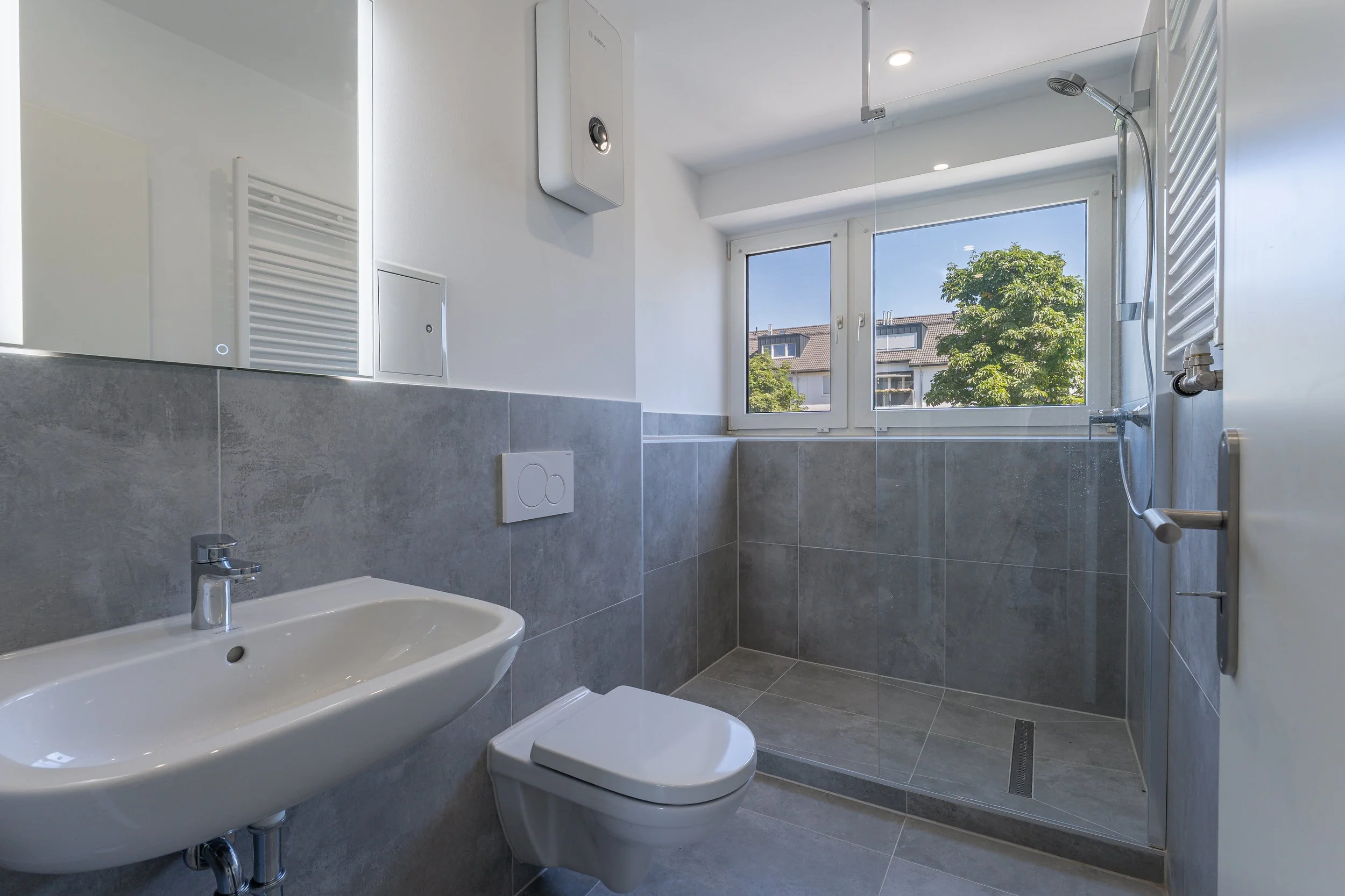 Badezimmer mit Waschbecken, Toilette, großzügiger Dusche mit Fenster, graue Fliesen, weiße Wände, moderne Ausstattung.