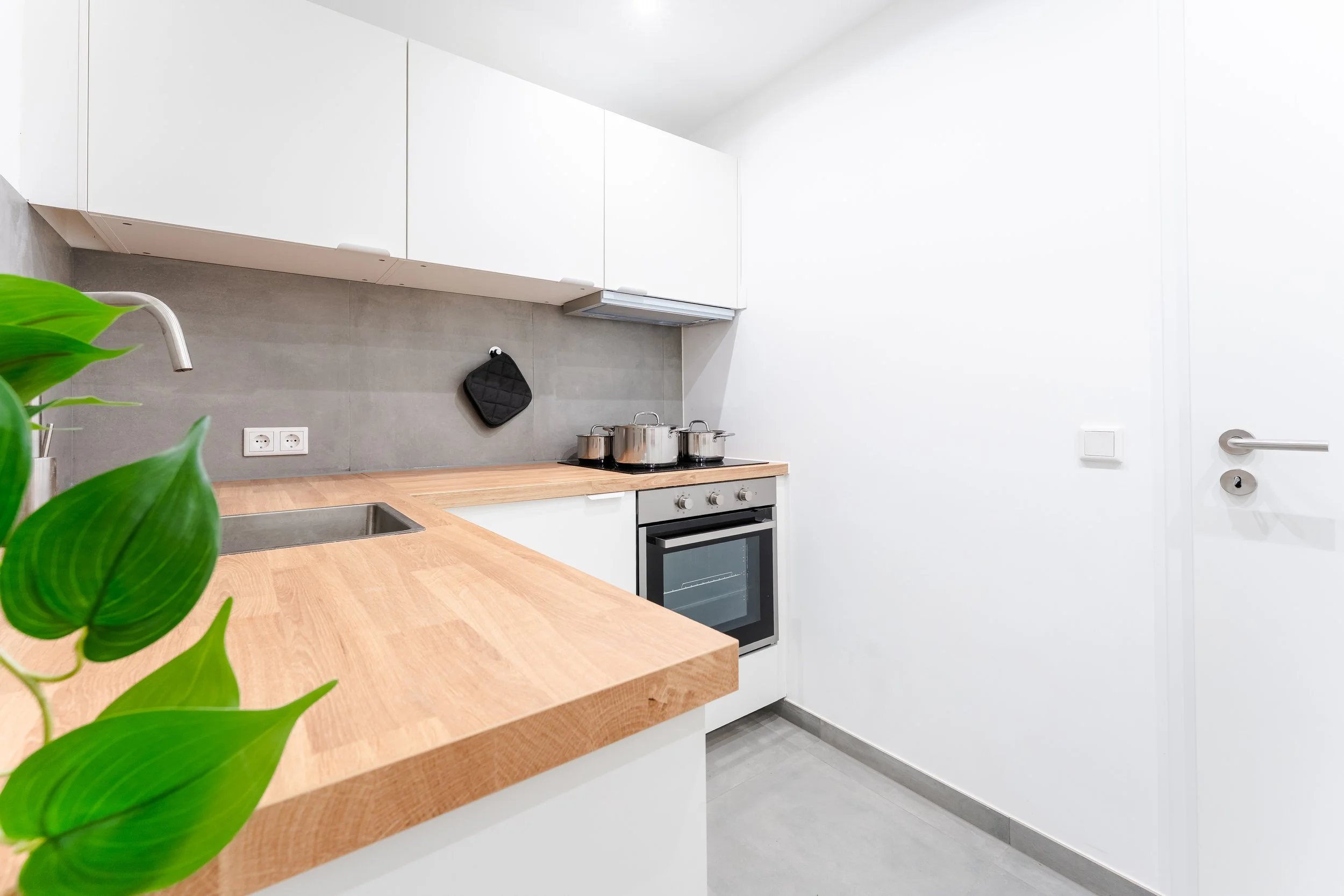Moderne helle Küche mit weißen Schränken, grauem Fliesenspiegel, Holzarbeitsplatte, Spüle, Elektroherd mit Töpfen, schwarzem Ofen, einer schwarzen Topflappen und einem grünen Pflanzenblatt im Vordergrund.