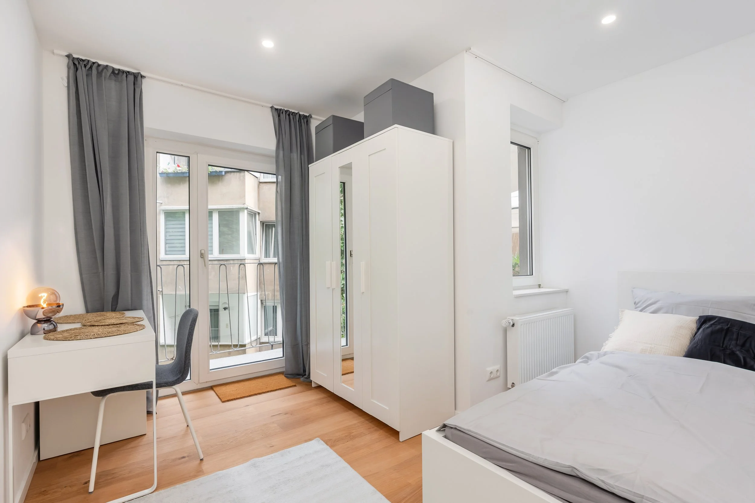 Schlafzimmer mit weißem Bett, Kleiderschrank, Fenster, Balkon mit Vorhängen, Holzboden.