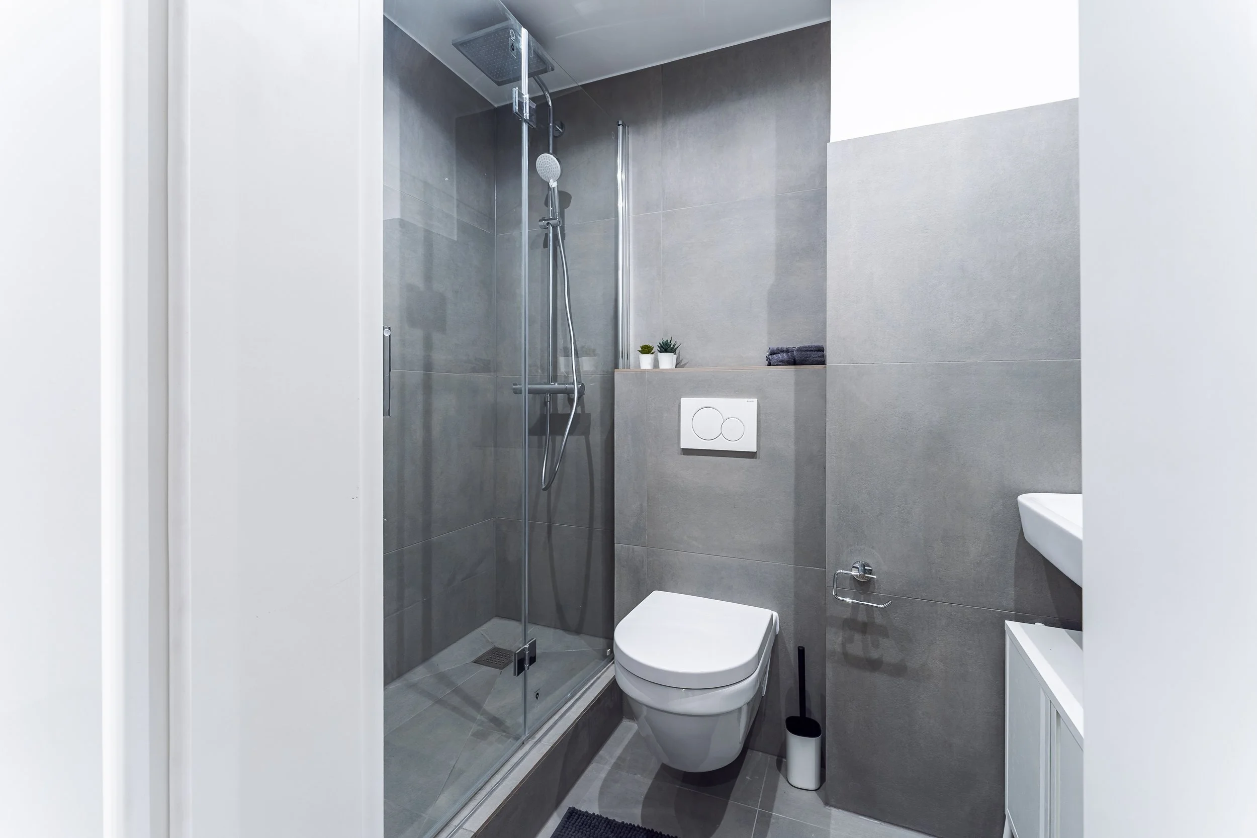 Modernes Badezimmer mit begehbarer Dusche, wandhängendem WC, kleines Waschbecken, grauen Fliesen und weißen Wänden, Dekoration in Form von kleinen Pflanzen.