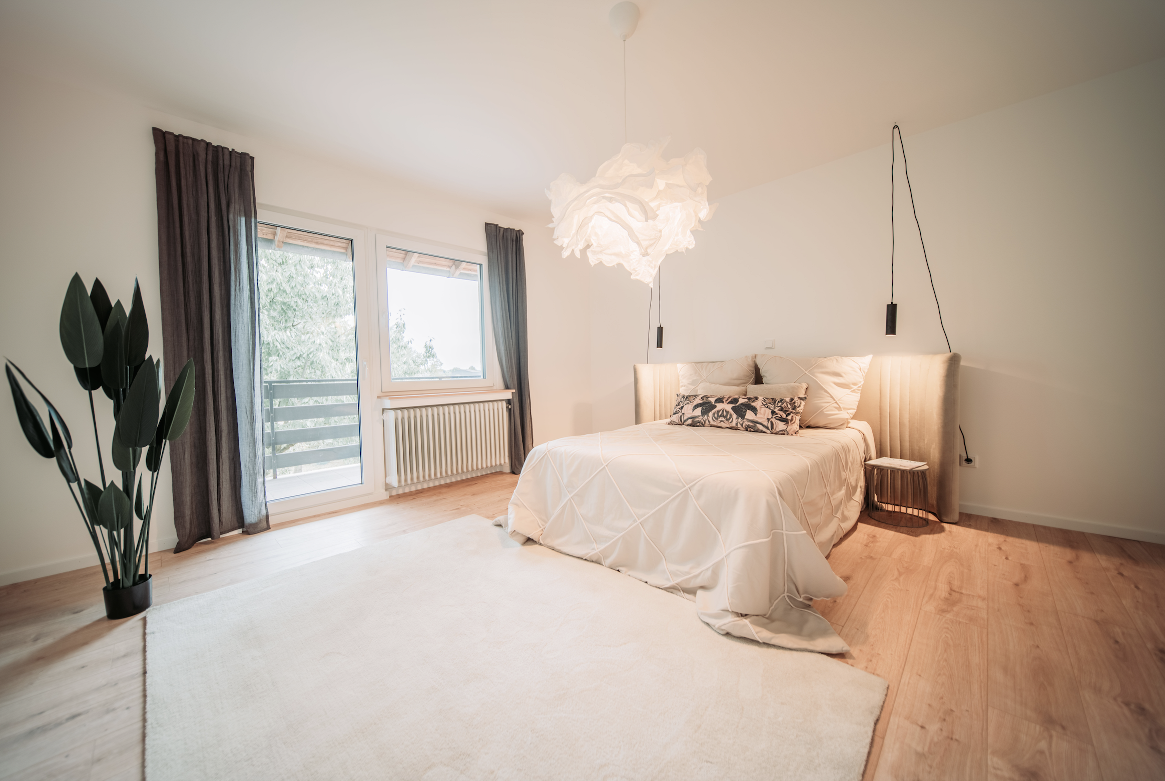 Gemütliches Schlafzimmer mit einem großen Bett, Nachttischen, einem hängelampenschirm, großen Fenstern mit dunklen Vorhängen, Teakholzboden und einer weißen Wand. Ein kleiner Stehtisch steht neben dem Bett, und es gibt eine Zimmerpflanze in einer sch