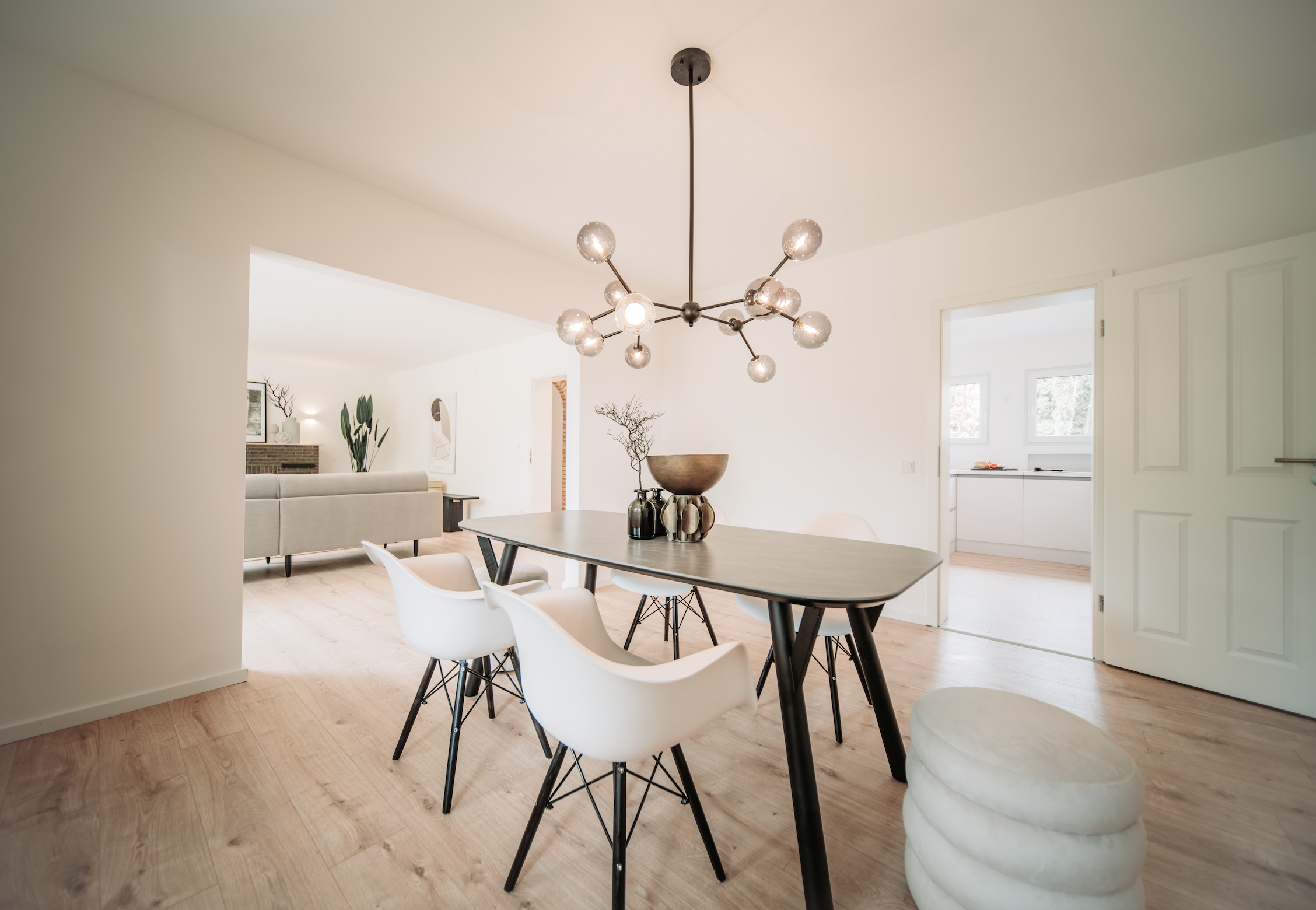 Moderner Esstisch mit weißen Stühlen, Dekokugeln und einer Tischlampe, offener Wohnbereich mit Sofa und Pflanzen, helle Wände, Holzboden, große Fenster im Hintergrund.