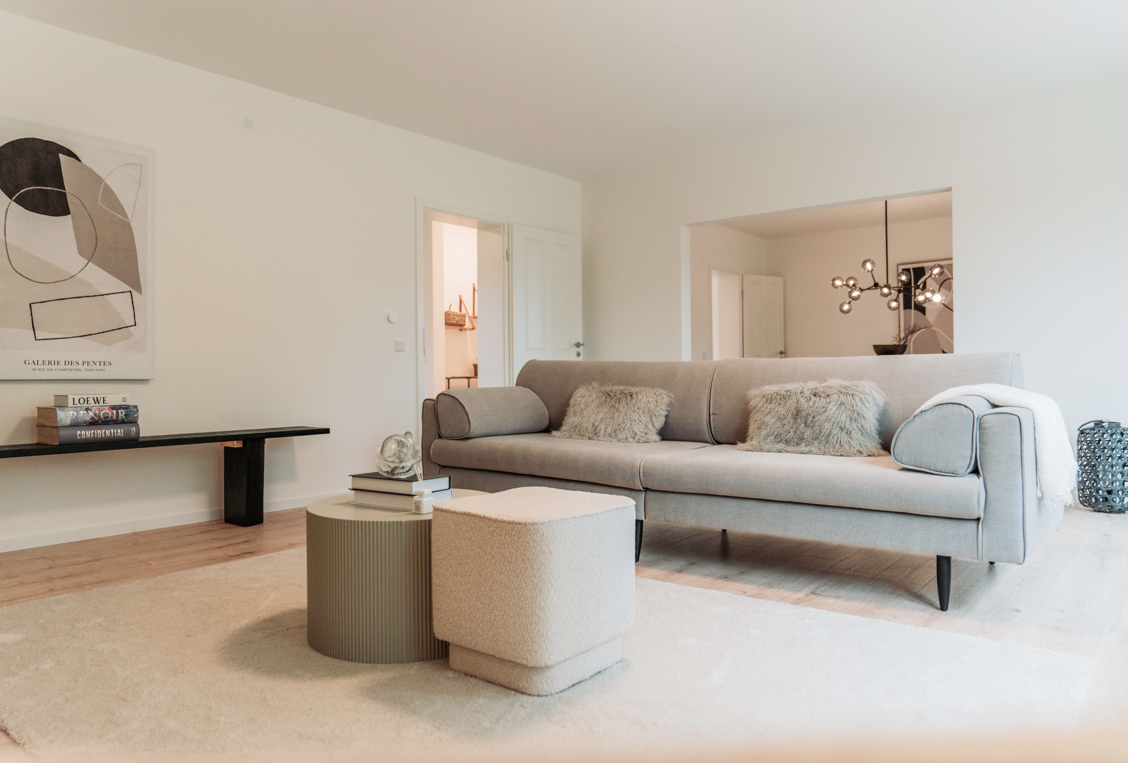 Modernes Wohnzimmer mit grauem Sofa, vier flauschigen Kissen, zwei quadratischen Beistelltischen, Wandkunst, offener Raum mit Beleuchtung, Dekorationsgegenständen und Teppich.