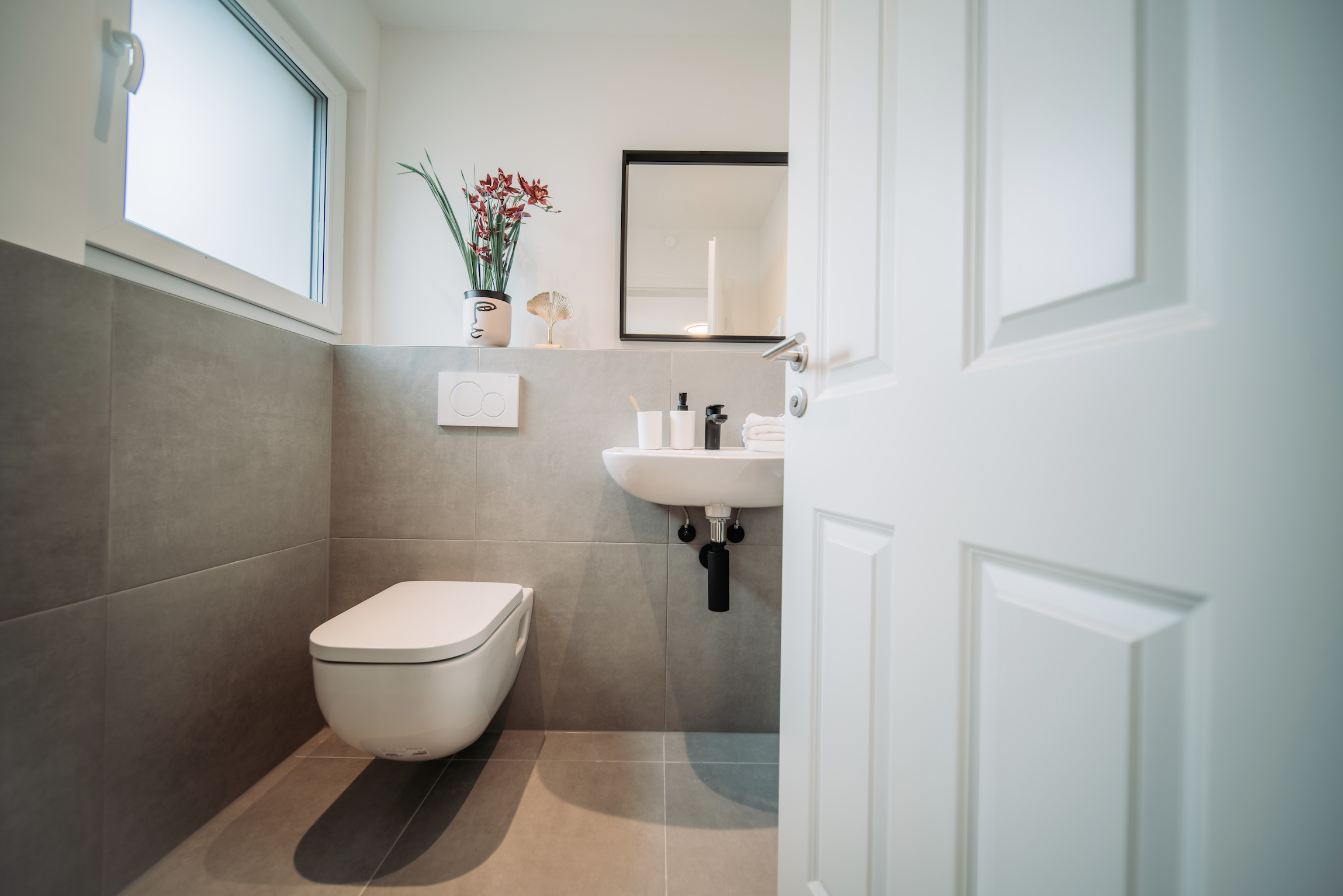 Ein modernes, kleines Badezimmer mit grauen Fliesen, einer weißen Toilette, einem kleinen weißen Waschbecken, einem Spiegel, einer Fenster mit Milchglas, einer Pflanze und Dekorationsgegenständen.