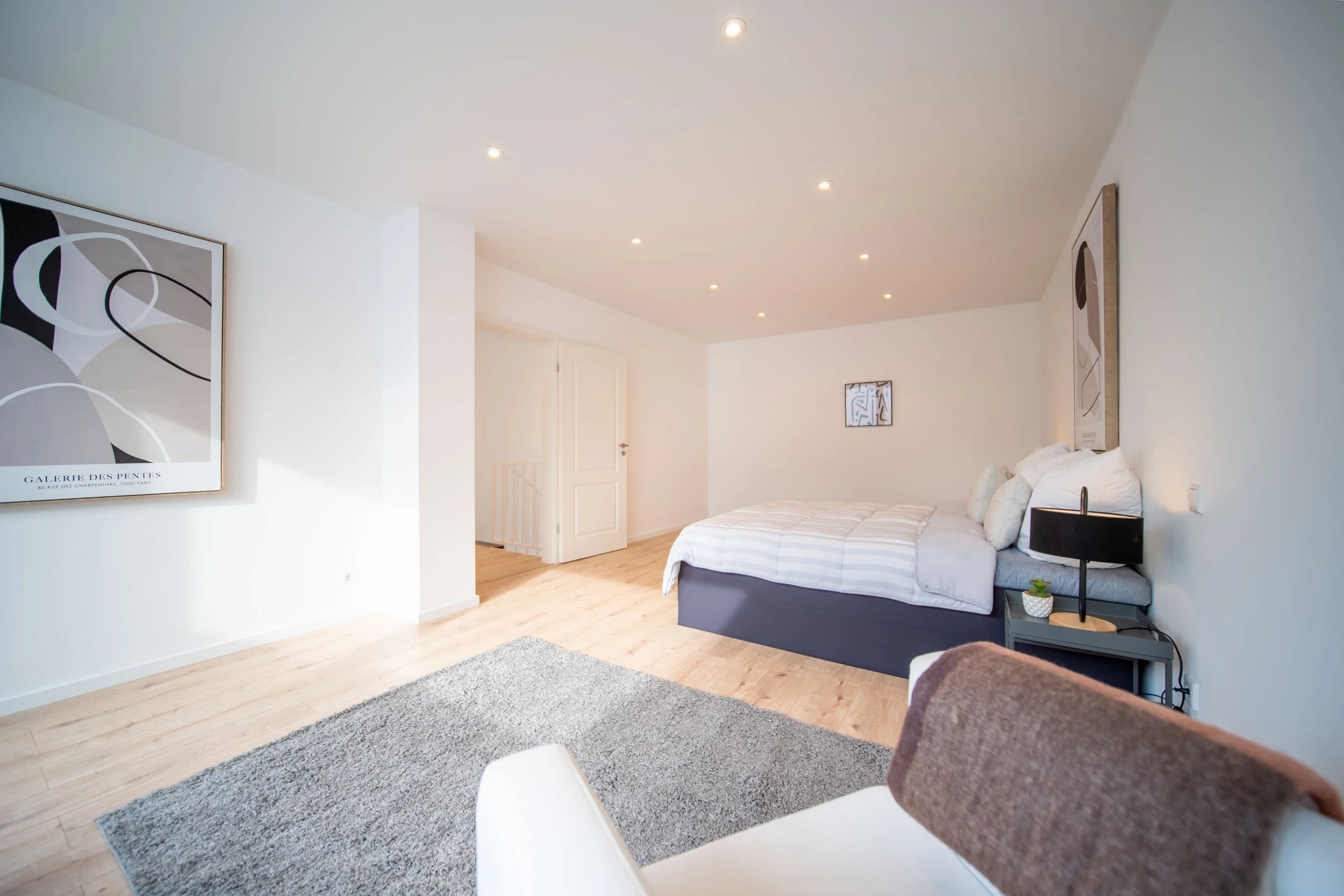 Ein modernes Schlafzimmer mit weißem Bett, grauem Nachttisch, schwarzer Lampe, abstrakten Wandkunstwerken, Holzboden, grauem Teppich und weißen Wänden.