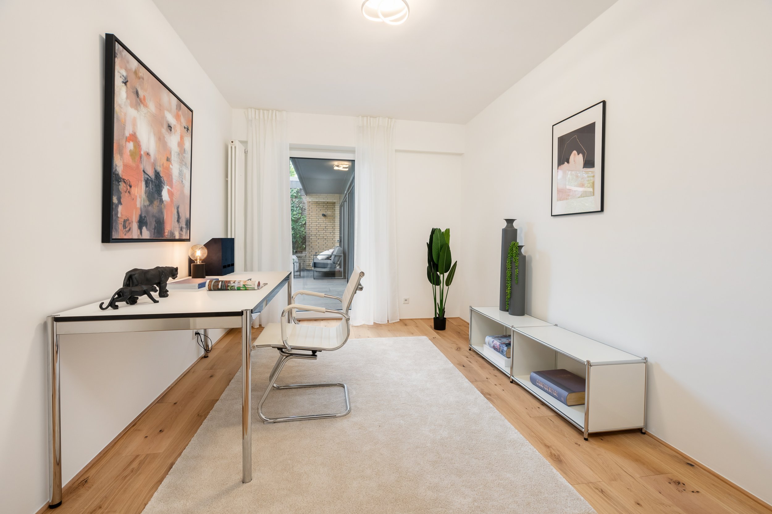 Modernes, minimalistisches Wohnzimmer mit weißen Wänden, Holzfußboden, einem weißen Teppich, einem weißen Schreibtisch mit schwarzen Elementen, einem modernen weißen Bürostuhl, einer Pflanzenvase, und einer Lowboard mit Dekorvasen und Büchern, Blick 