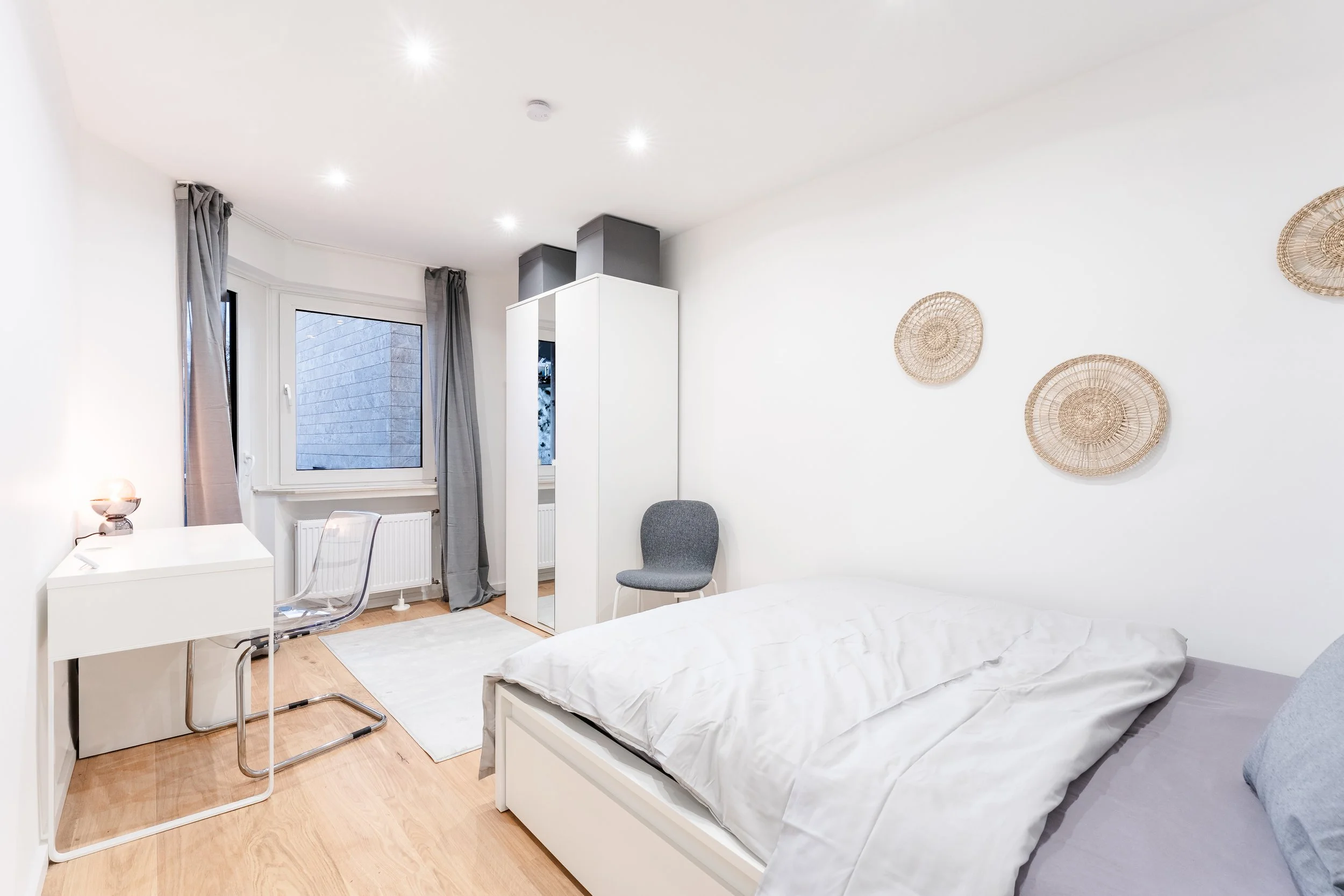 Schlafzimmer mit weißem Bett, grauen Kissen, weißen Wänden, Wanduhr aus Bast, Fenster mit grauen Vorhängen, kleiner Schreibtisch mit transparenter Stuhl, weiterer grauer Stuhl, weißer Schrank, Holzboden, kleine Stehlampe auf der Kommode.
