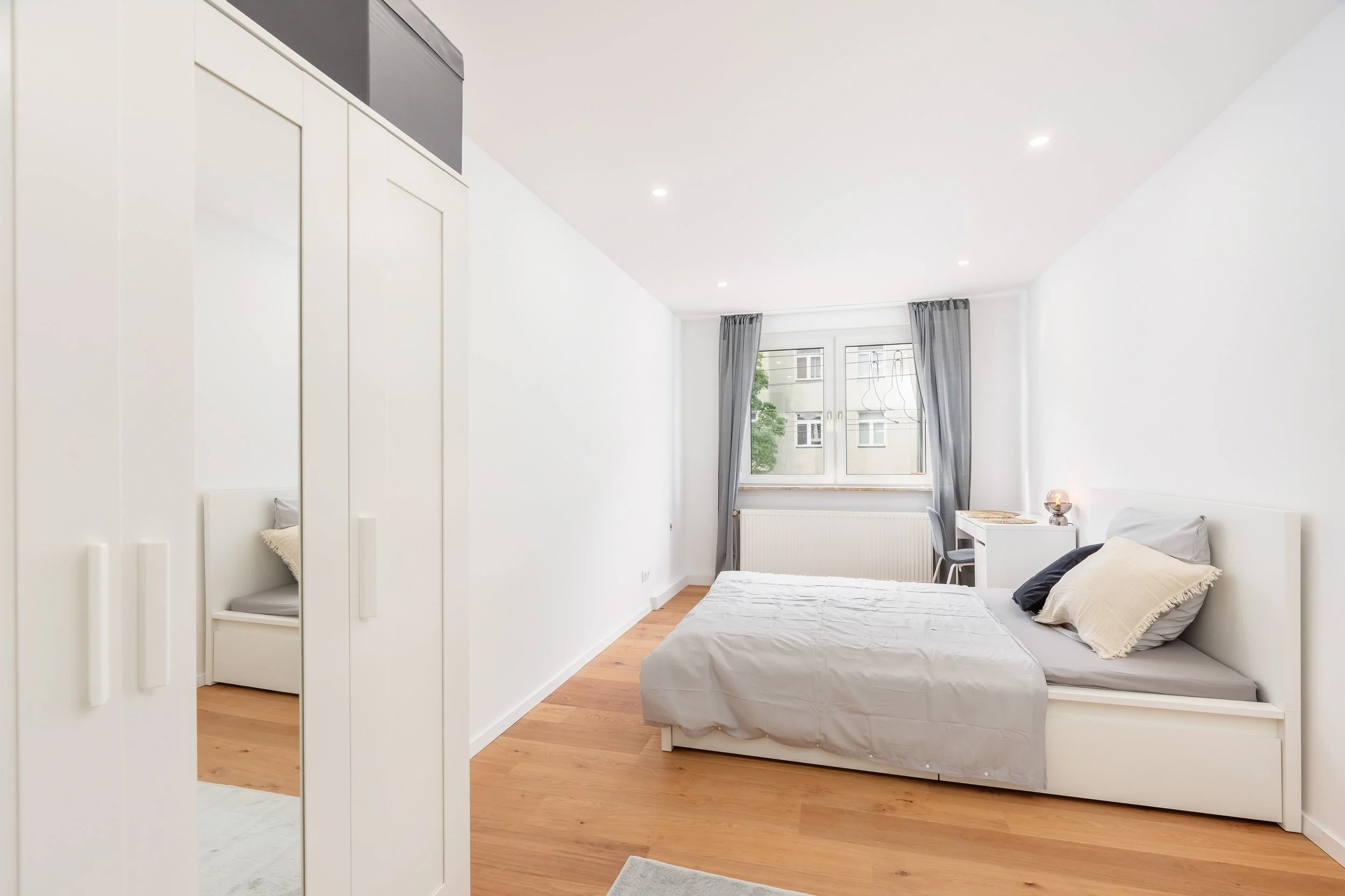 Schlafzimmer mit weißen Möbeln, Holzfußboden, Bett mit grauer Bettwäsche und Kissen, großer Fenster mit grauen Vorhängen, kleine Schreibtisch mit Stuhl, Nachttisch mit Lampe, minimalistisch gestaltet.