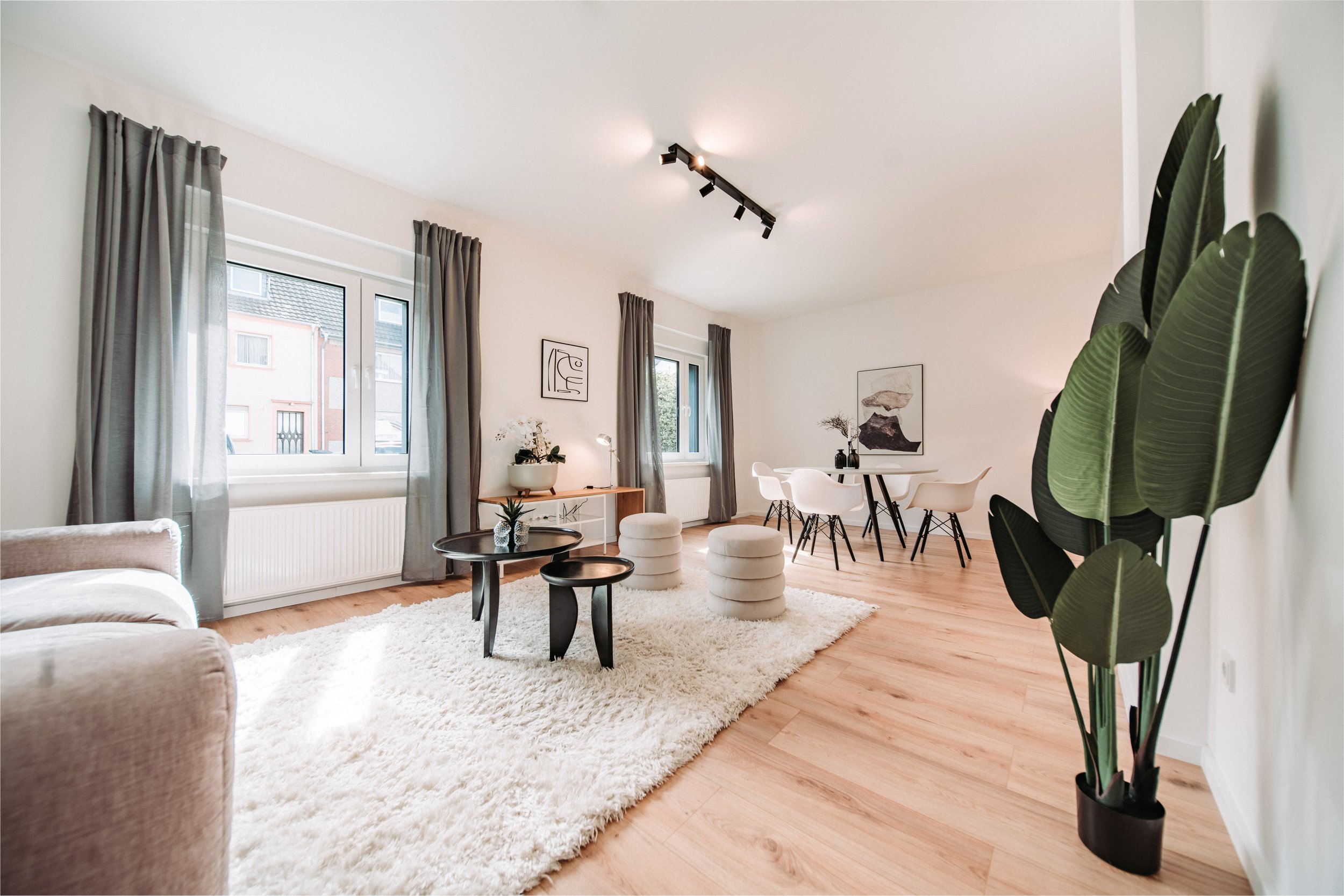 Modernes Wohnzimmer mit weißen Wänden, Eichenholzboden, grauen Vorhängen, einer großen Fensterfront, einem weißen Sofa, einem schwarzen Couchtisch, einer weißen Teppich, mehreren Sitzhockern, einem Essbereich mit weißen Stühlen und einem runden Tisch