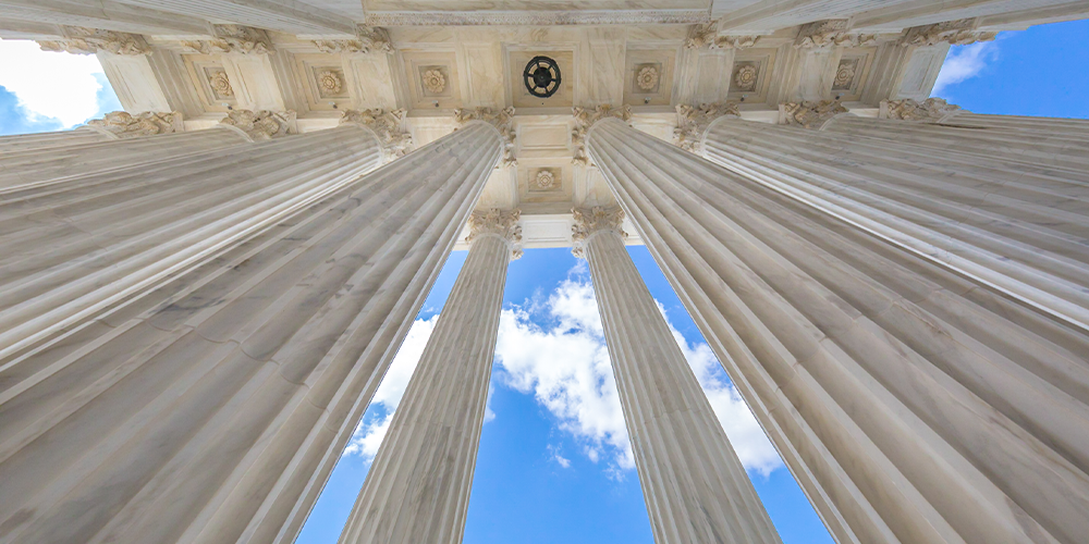 Looking up at classical columns supporting a decorated entablature against a blue sky with clouds.