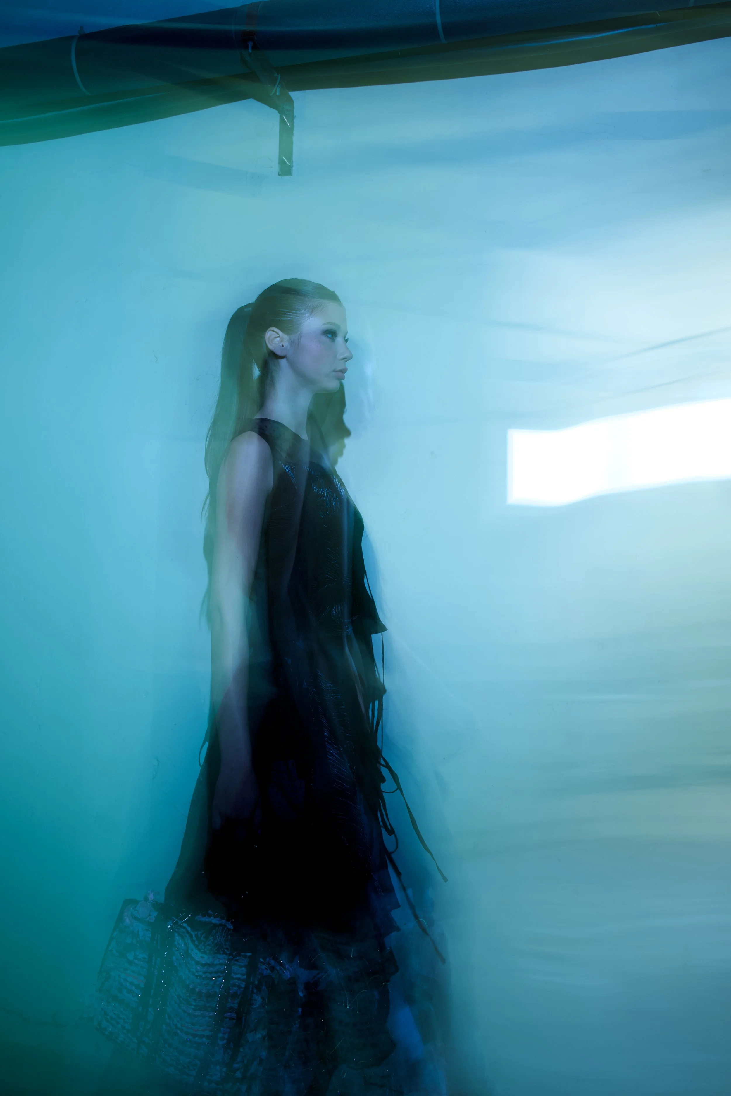 A woman standing sideways with reflection overlays in a blue-green space, holding a patterned bag, with a high-contrast lighting backdrop.