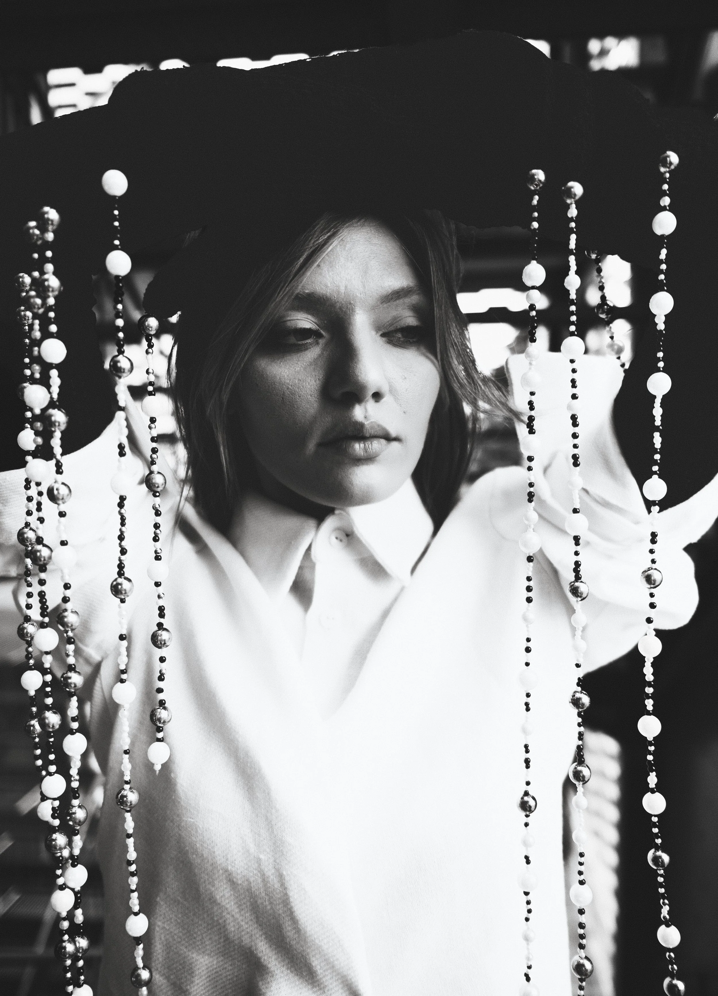 A woman with shoulder-length hair, wearing a white shirt, holding a large hat decorated with beads, in a black-and-white photograph.