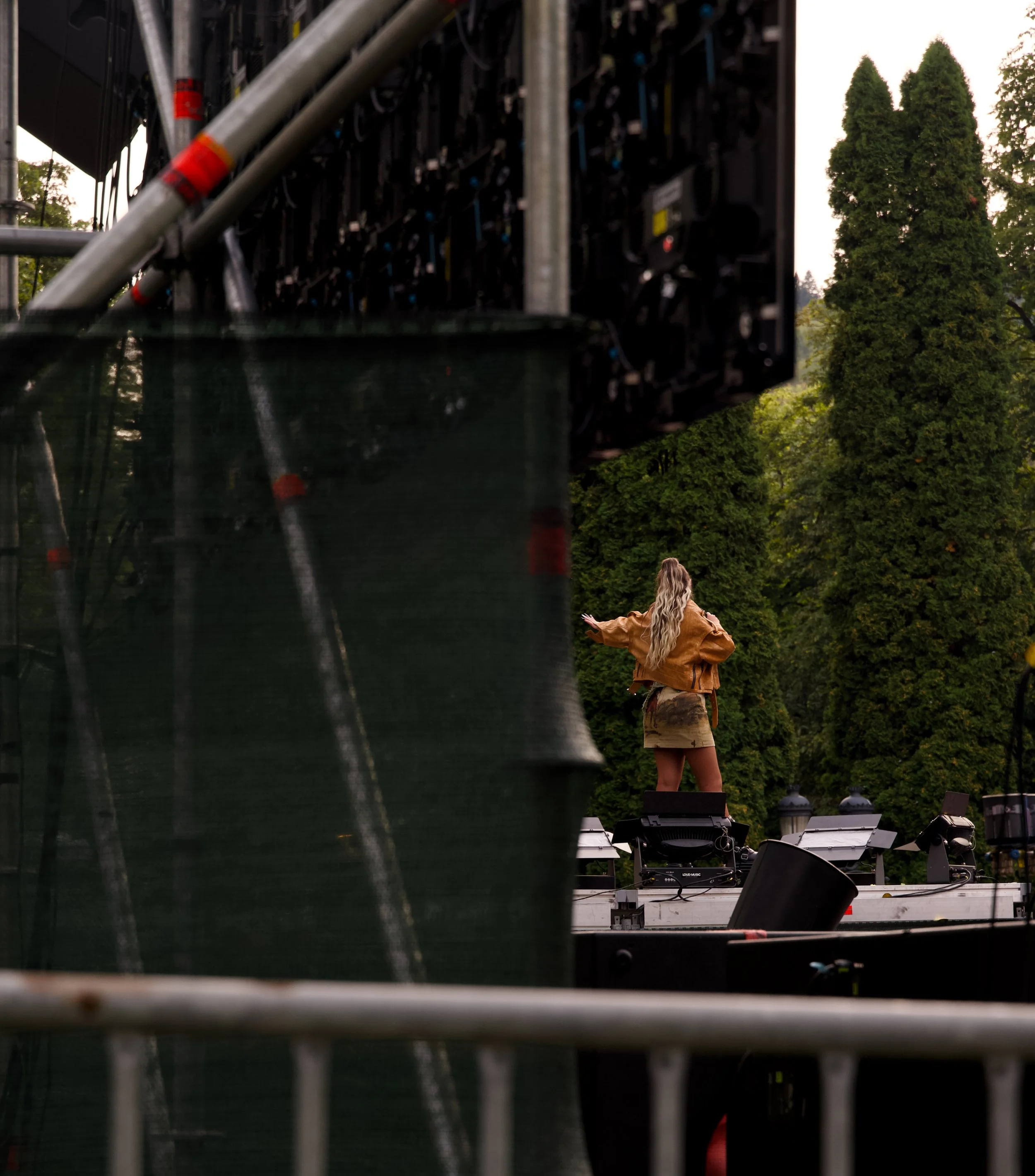 A woman with long blonde hair on stage in front of tall green trees, wearing a brown jacket and a short skirt, withStage and equipment visible.