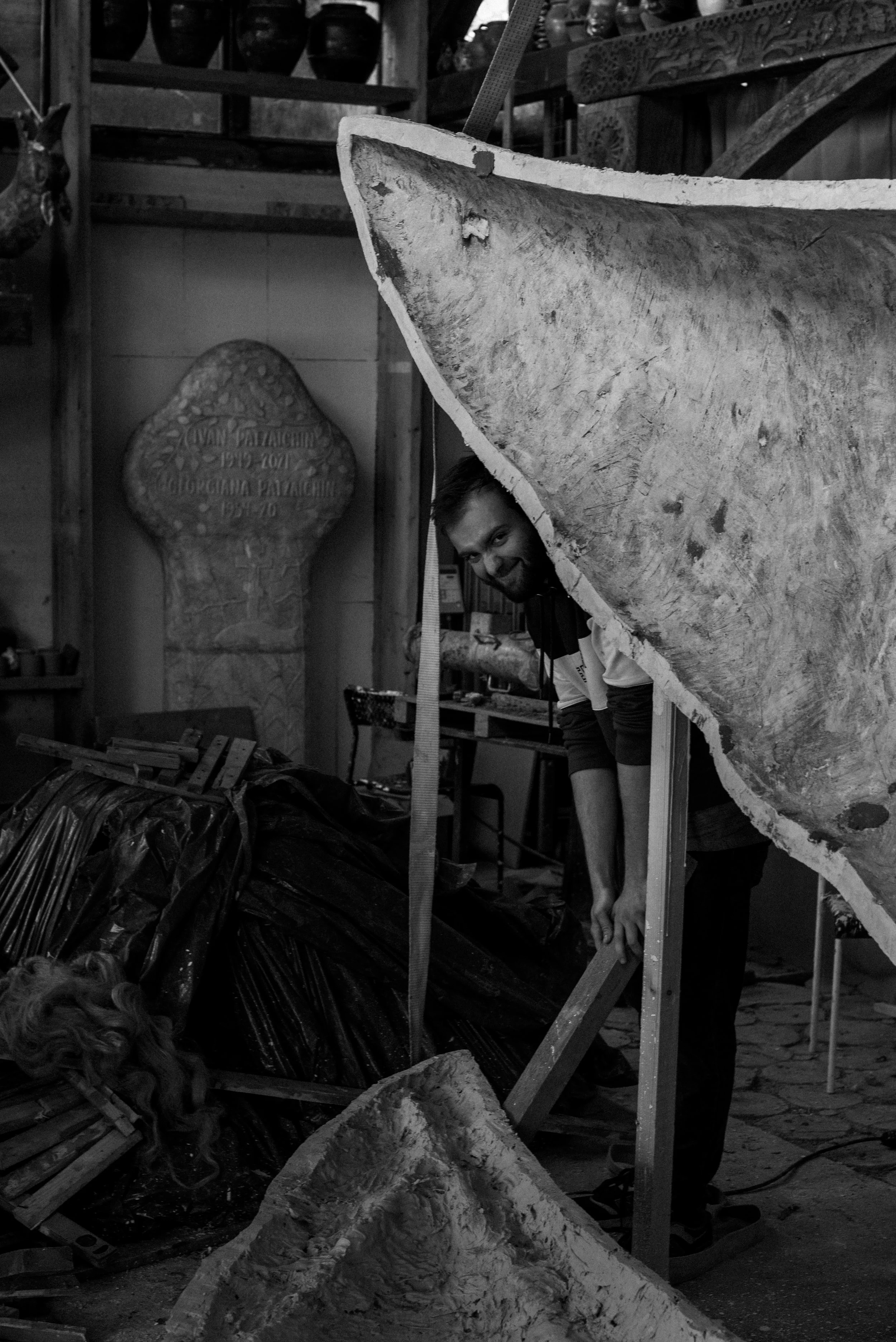 A man in a workshop peeking through a large, irregularly shaped piece of material, possibly stone or wood, with various tools and materials scattered around.