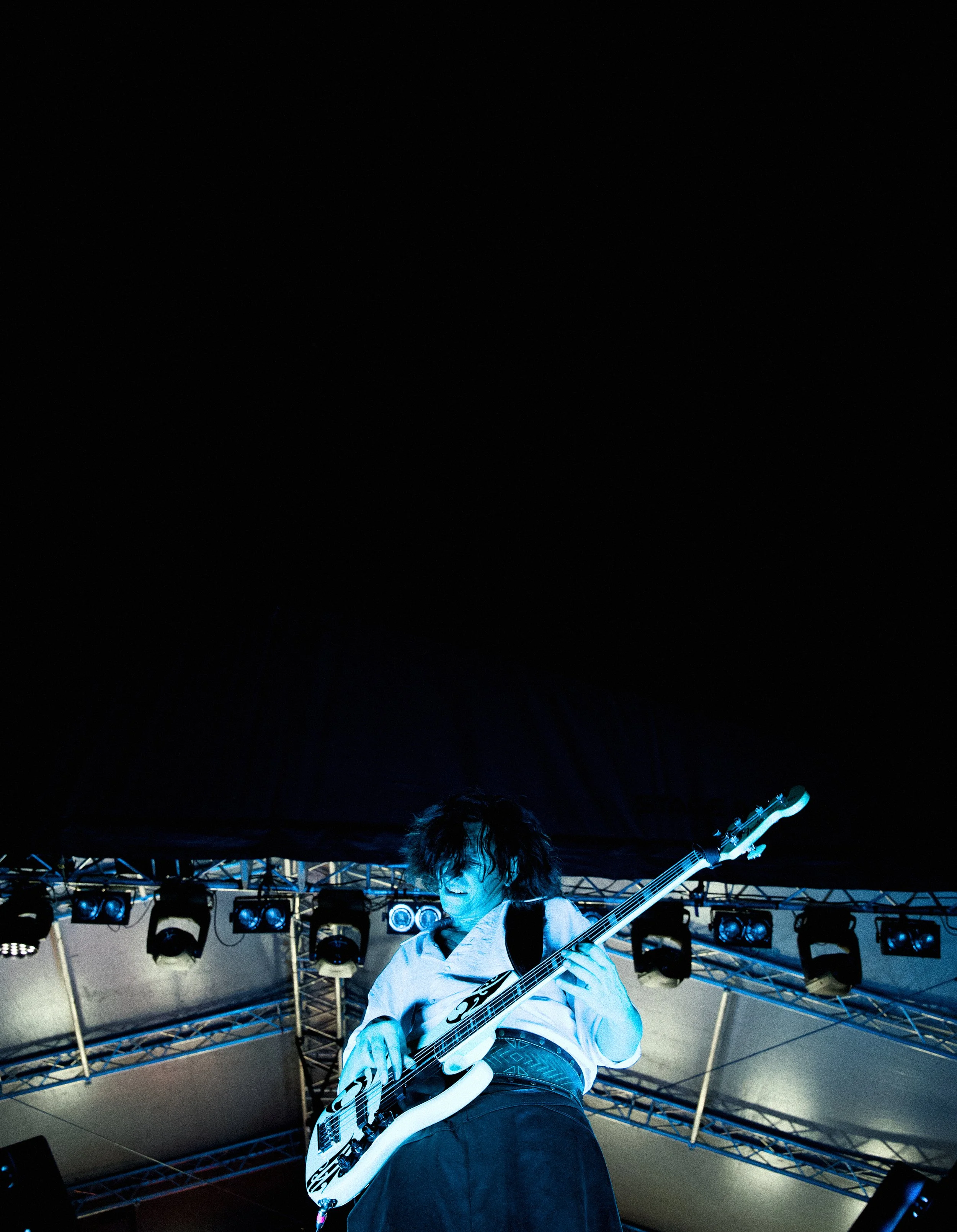 A person playing a white electric guitar on stage with stage lights and metal scaffolding in the background under a dark sky.