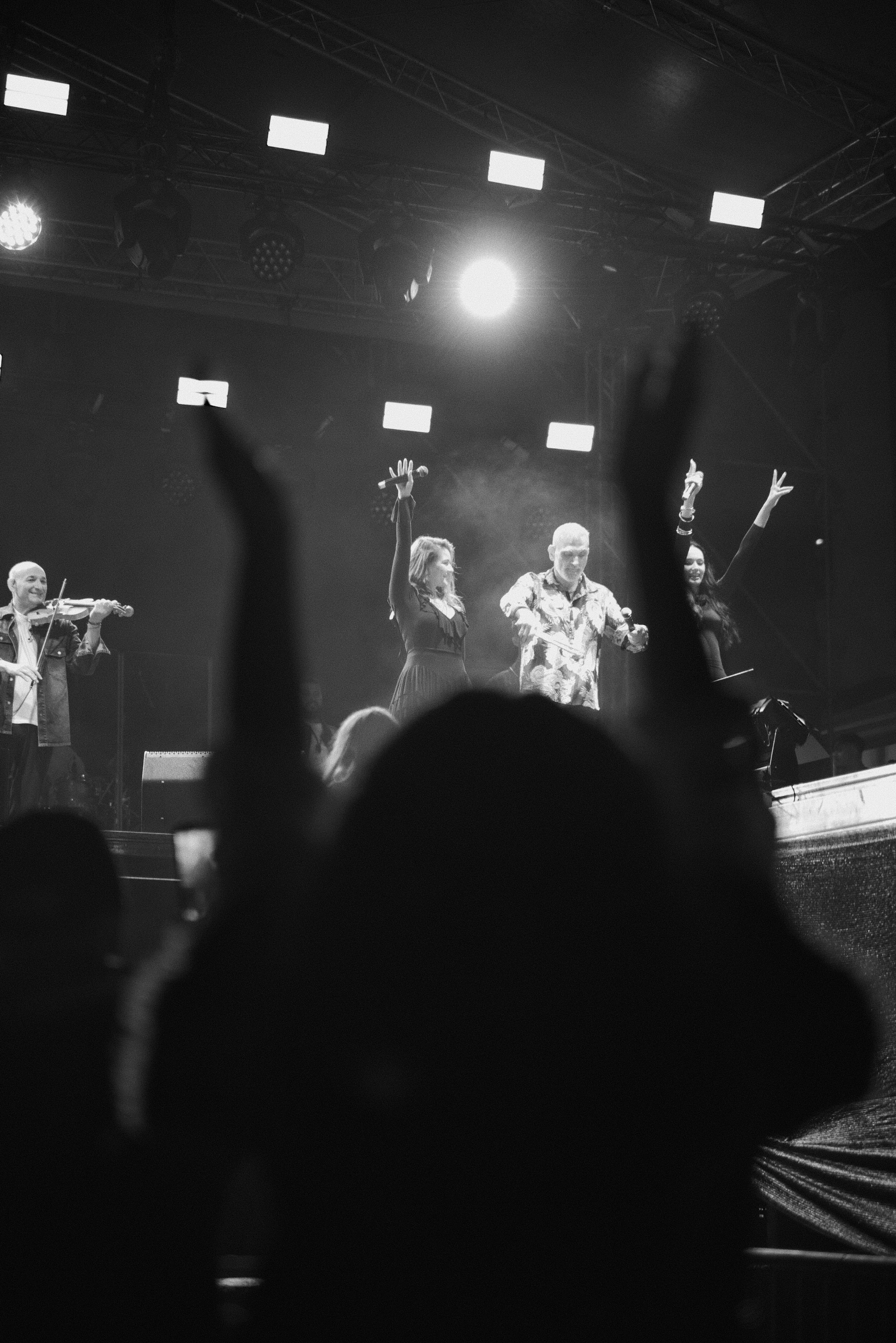 Black and white photo of a stage with four performers, two women and two men, singing and playing instruments; the audience's hands are raised in the foreground.