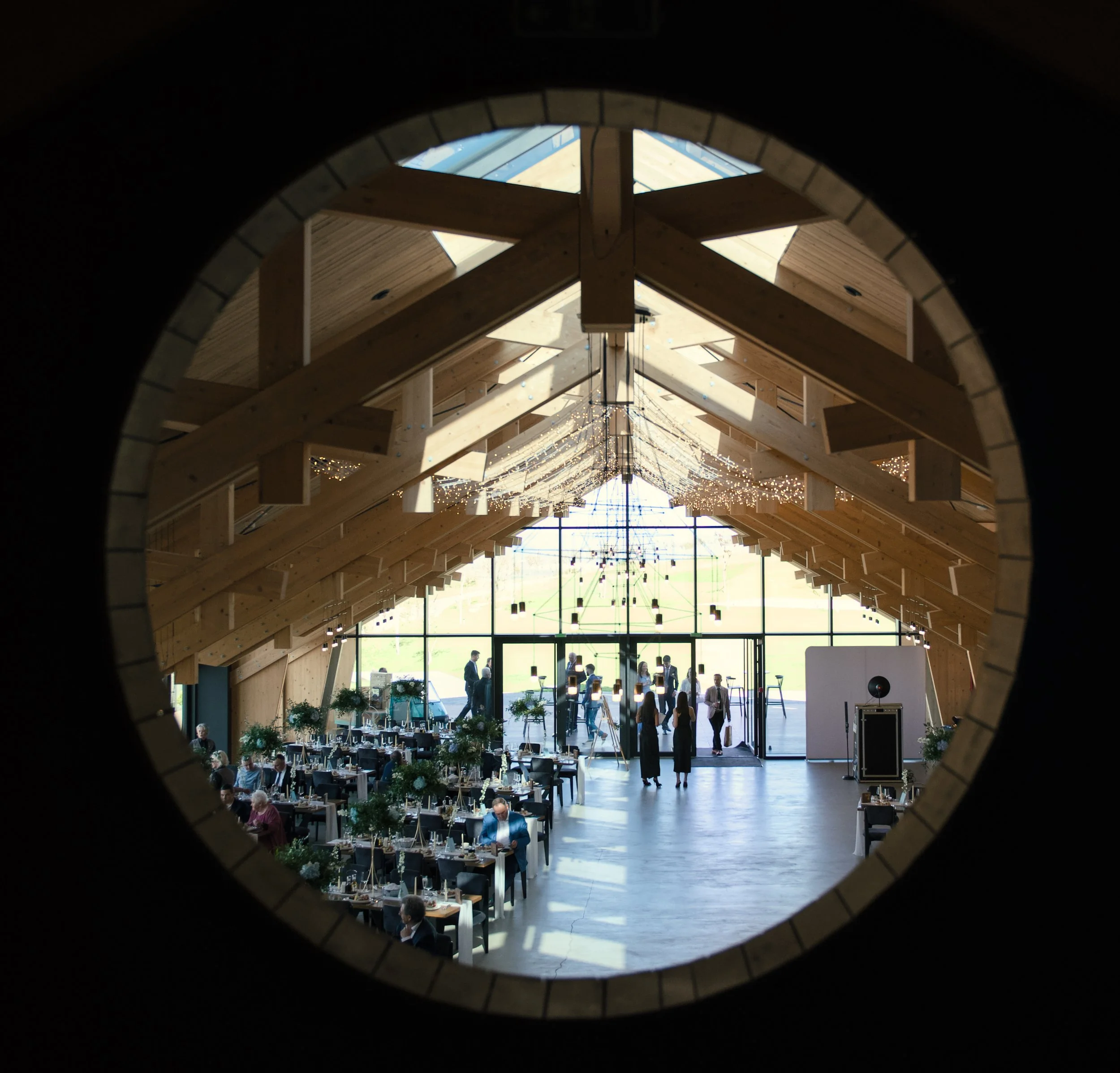 View of a modern event hall with wooden A-frame ceiling, large glass front, and people mingling inside during a celebration.