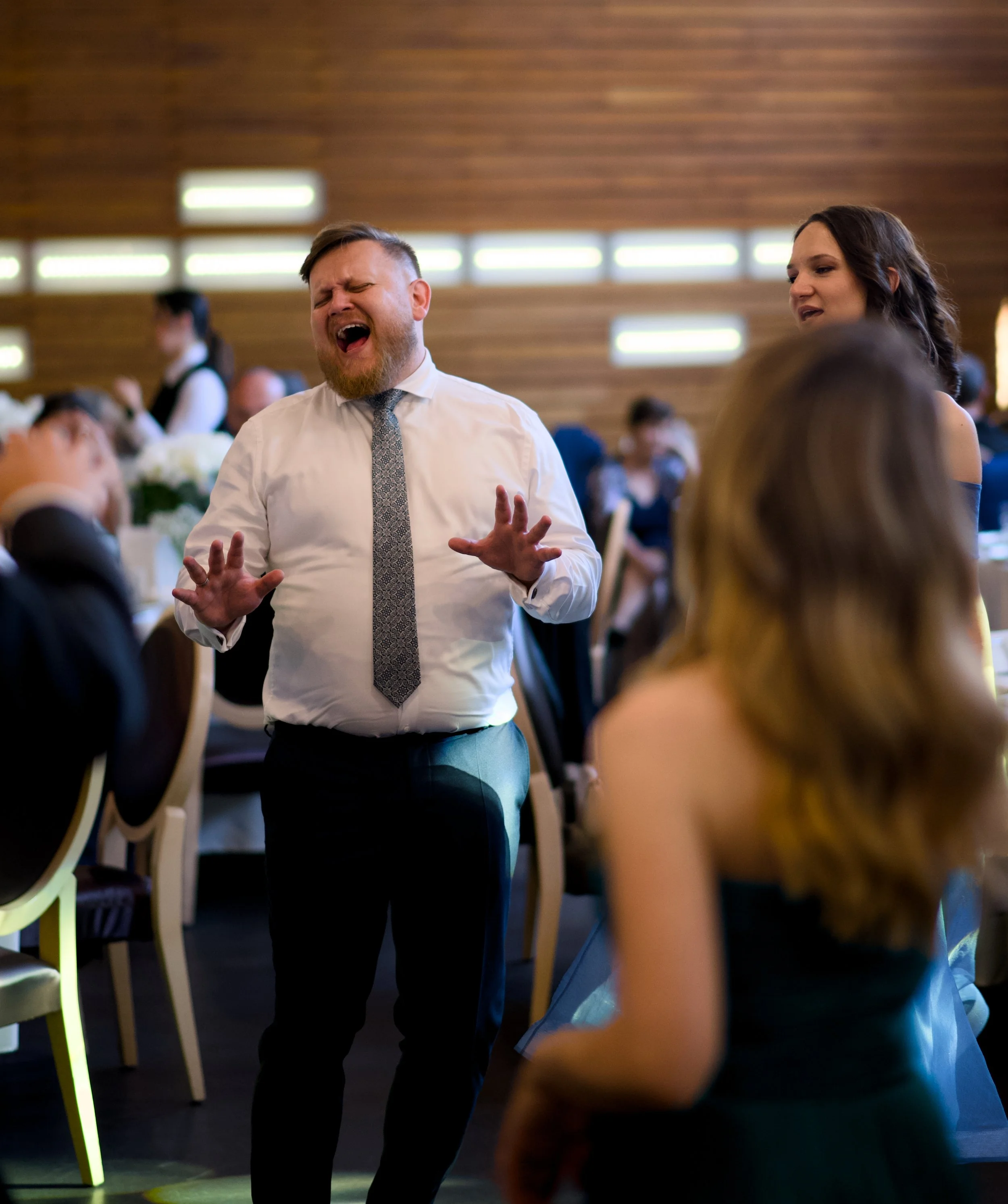 A man singing with his eyes closed and hands raised at a formal event, surrounded by seated guests, in a decorated venue with wood-paneled walls.