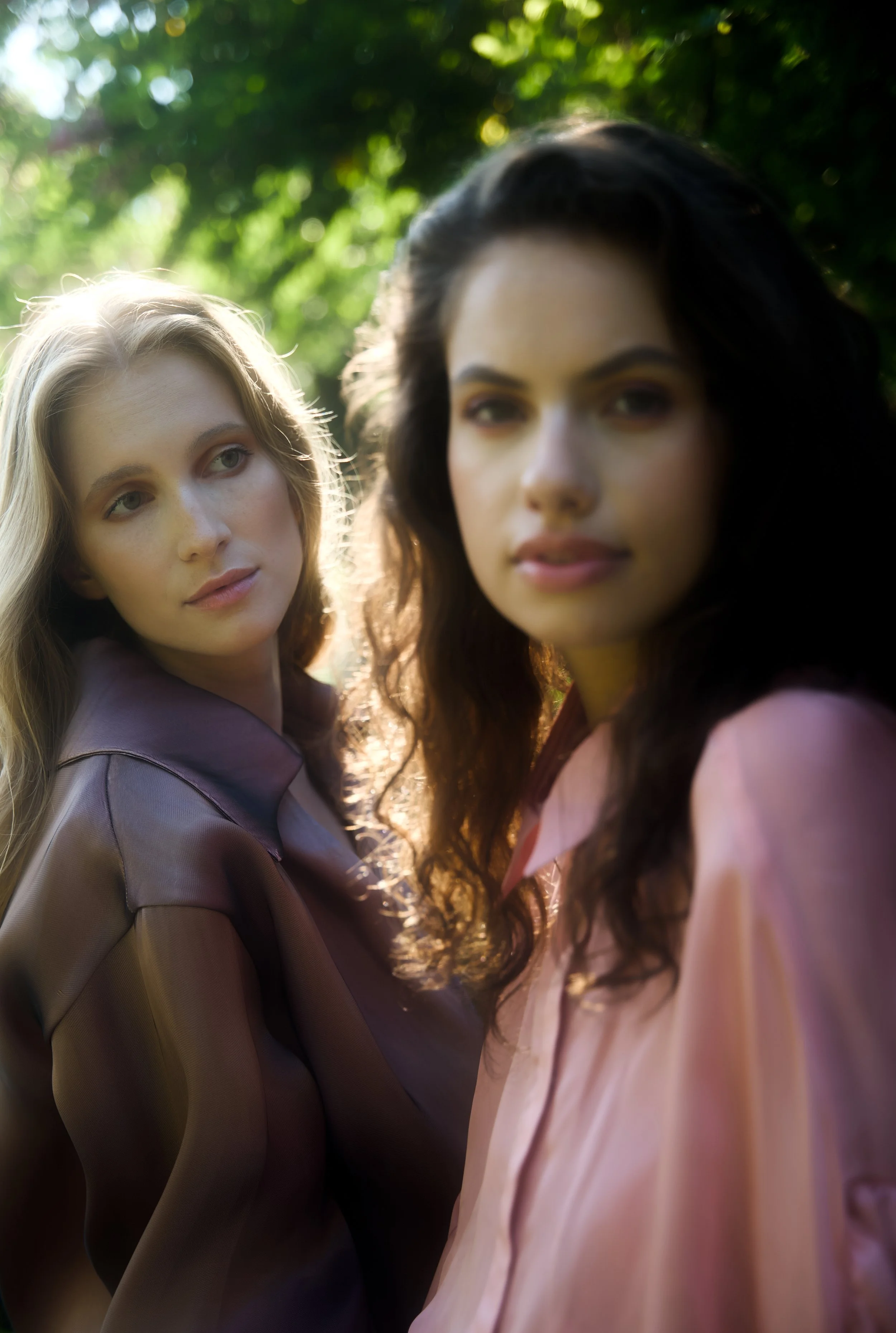 Two women outdoors, one with blonde hair and the other with dark curly hair, standing close in sunlight with greenery in the background.