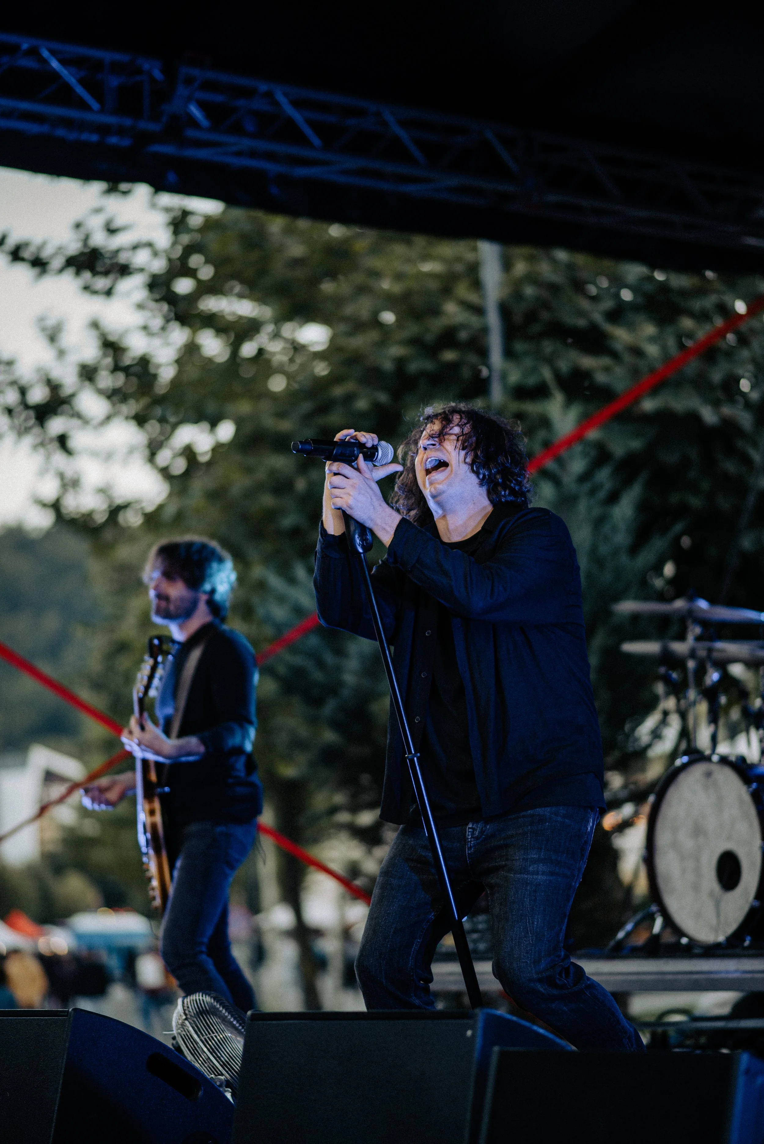 Musician singing into a microphone during an outdoor concert, with a guitarist in the background and a drum set nearby.