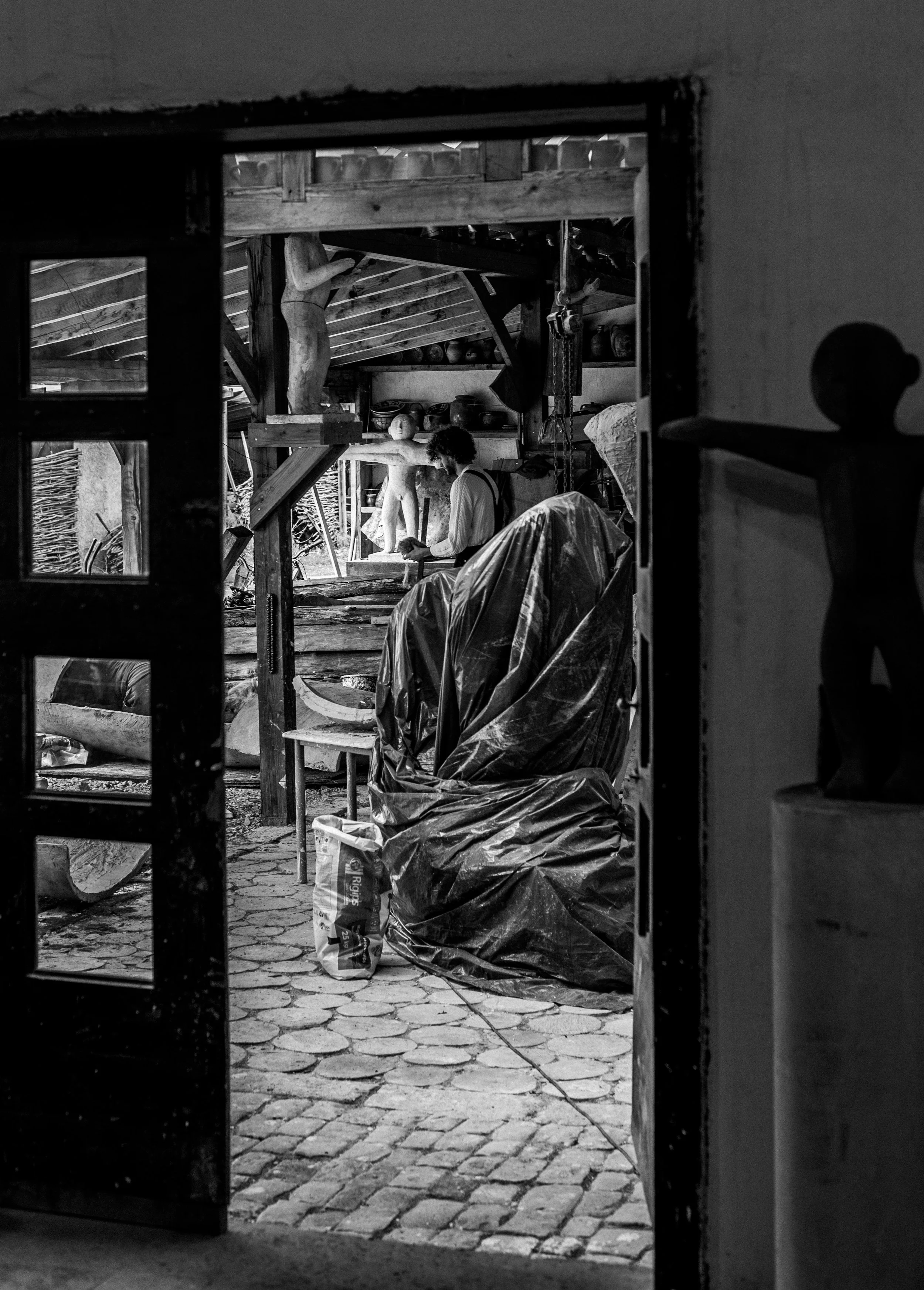 A black and white photograph of a woodworking workshop, viewed through a doorway. A man with curly hair is working on a wood sculpture of a human figure, with various tools and supplies around. The workshop has wooden beams and shelves filled with bo