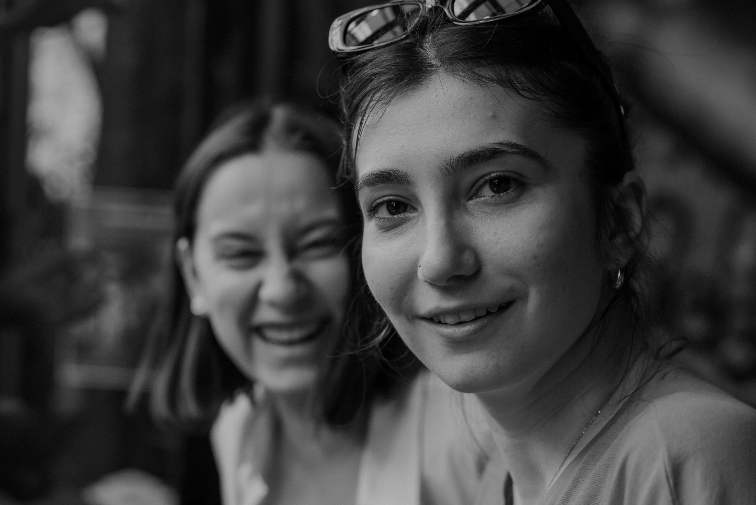 Two young women smiling, one in the foreground with sunglasses on her head and earrings, and the other in the background with glasses.
