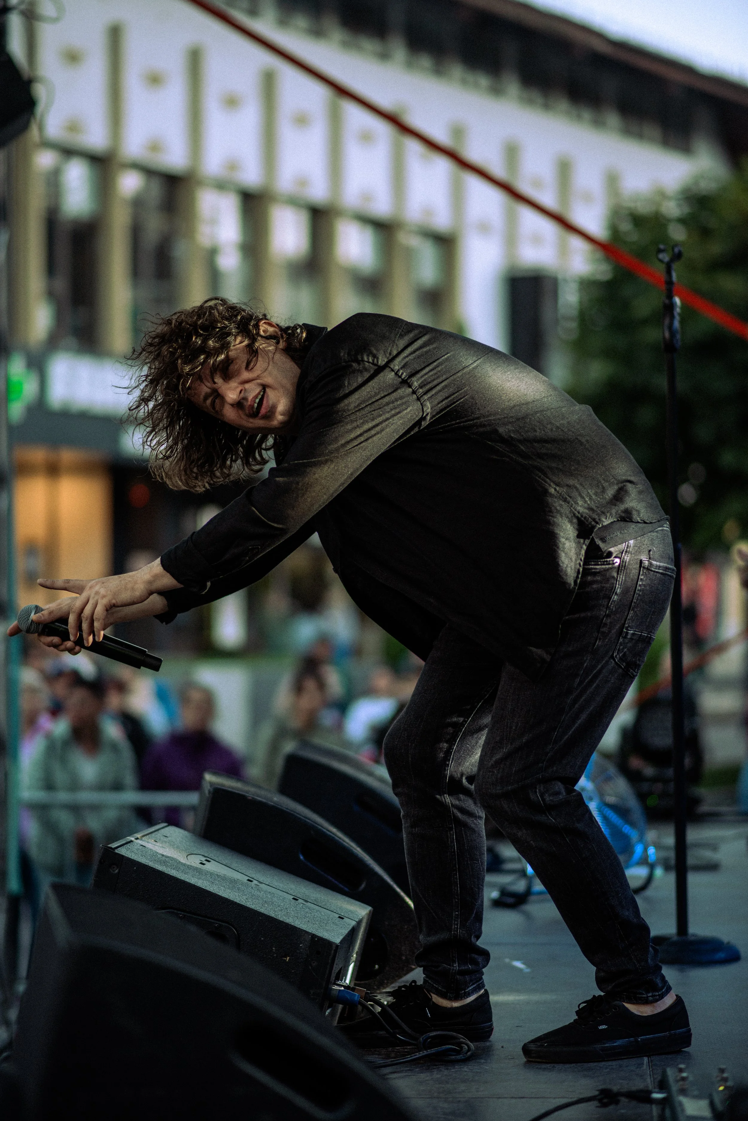 A man performing on an outdoor stage, leaning forward and holding a microphone, smiling and engaging with the audience.