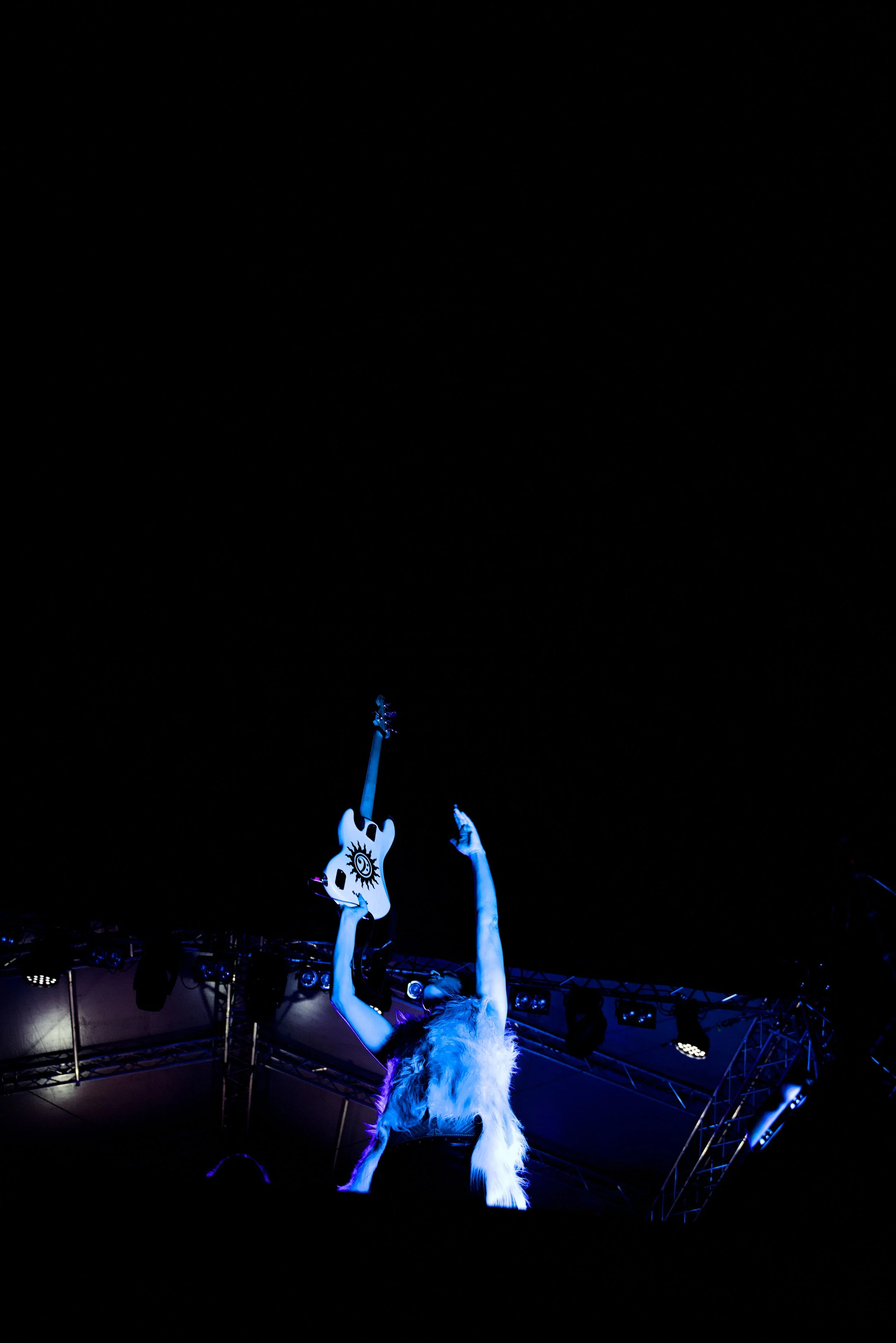 Performer on stage holding a white guitar with a black sunburst design, wearing a white fur coat and raising one arm, with stage lights and metal scaffolding in the background.