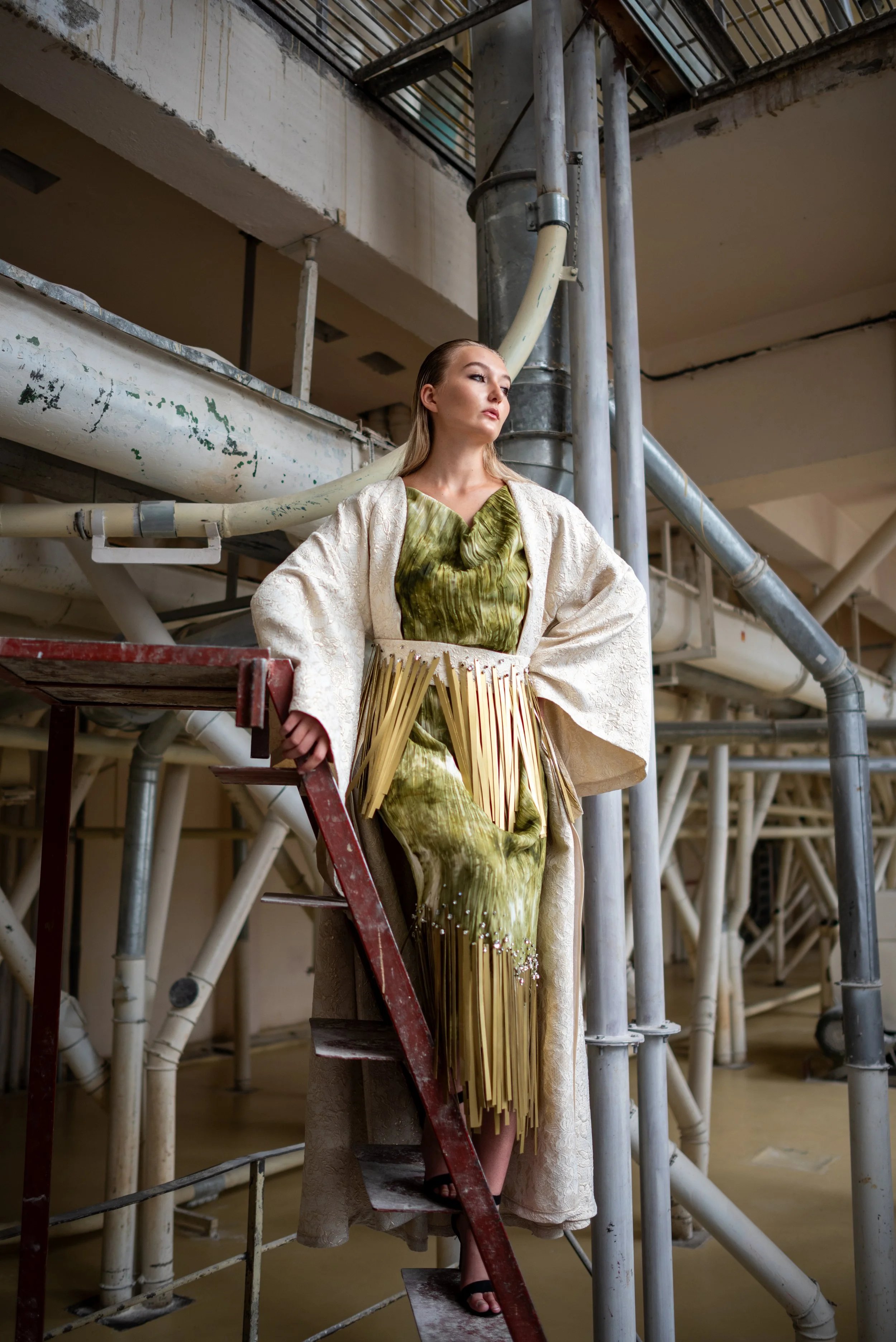 A woman in a creative green and white outfit, with an intricate skirt and textured top, stands amidst industrial pipes and scaffolding, leaning on a ladder.