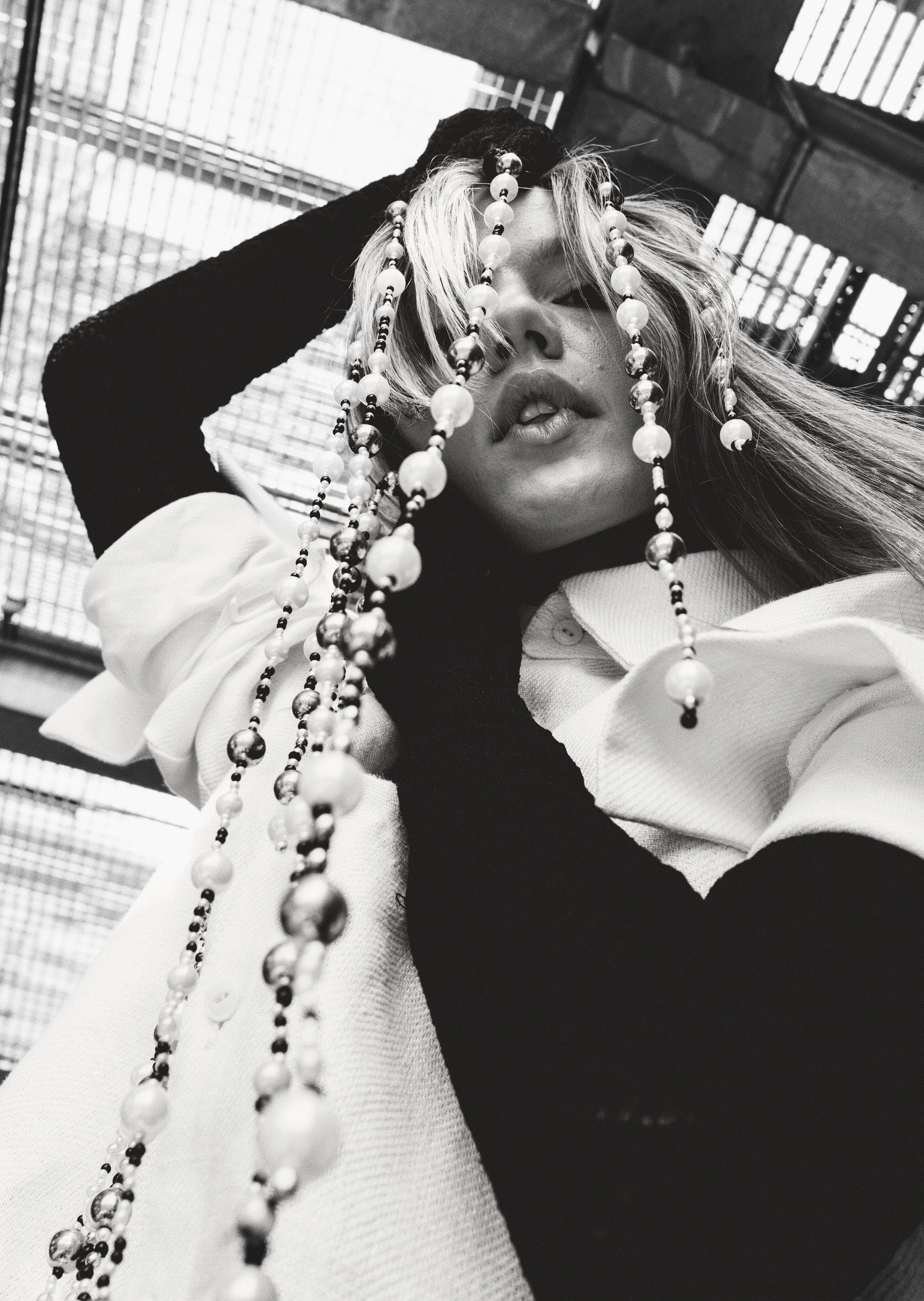 Black and white photograph of a woman with long hair, wearing a white shirt and holding strands of beads in front of her face, with a background of a metal and brick structure.