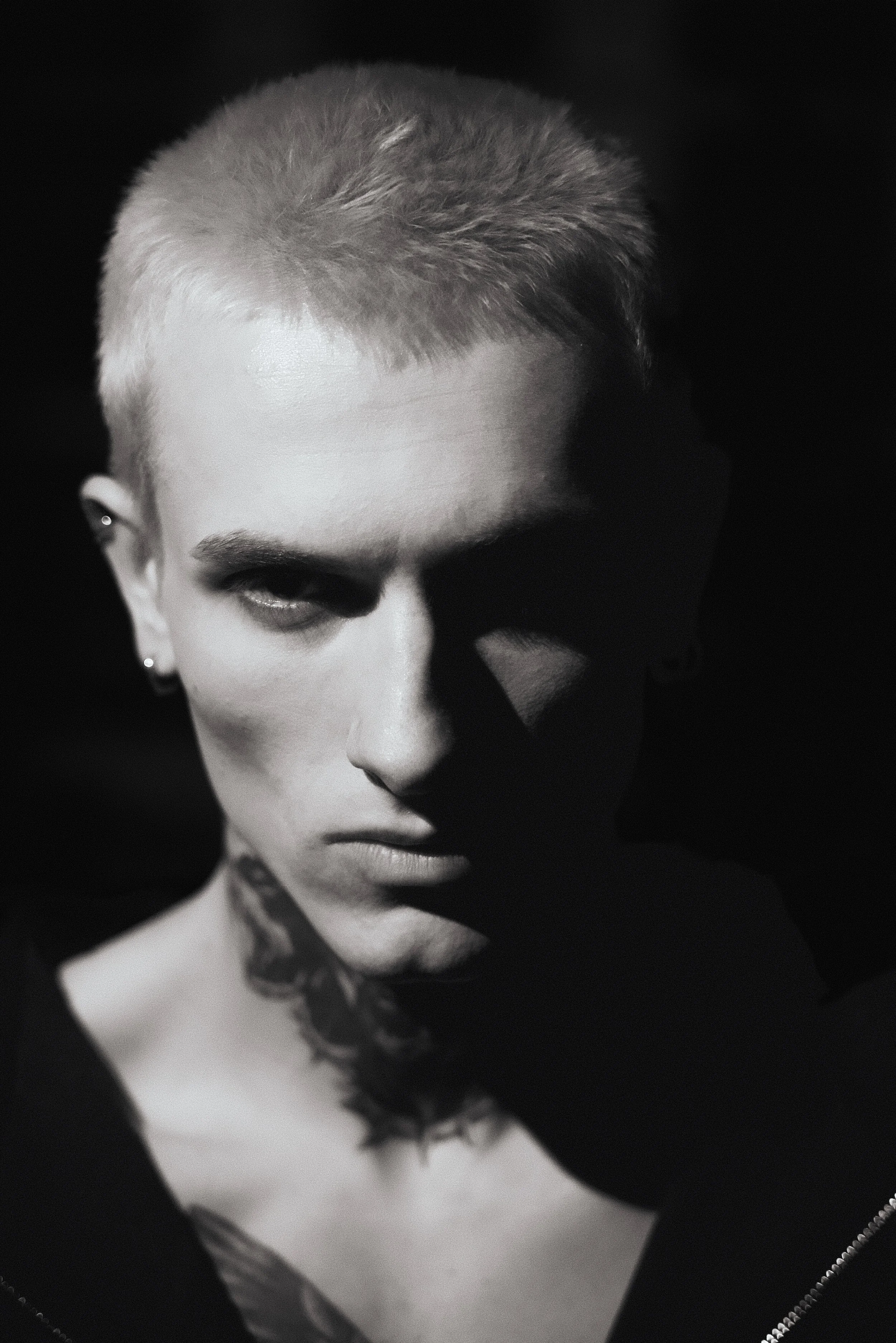 Black and white portrait of a person with short hair, earrings, and a tattoo on their neck, with dramatic lighting creating strong shadows on their face.