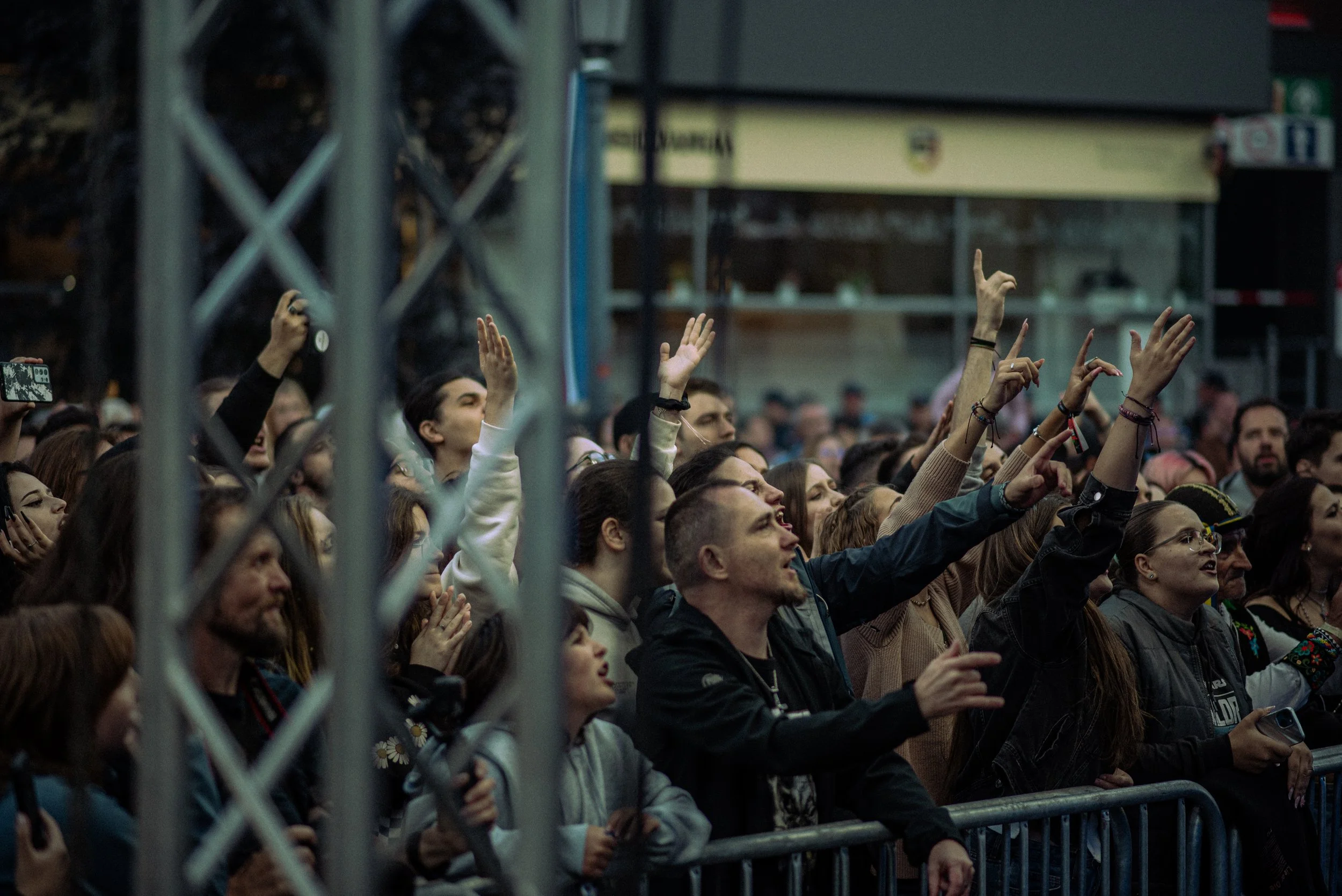 Crowd of people attending a concert, some raising their hands, behind a metal barrier.