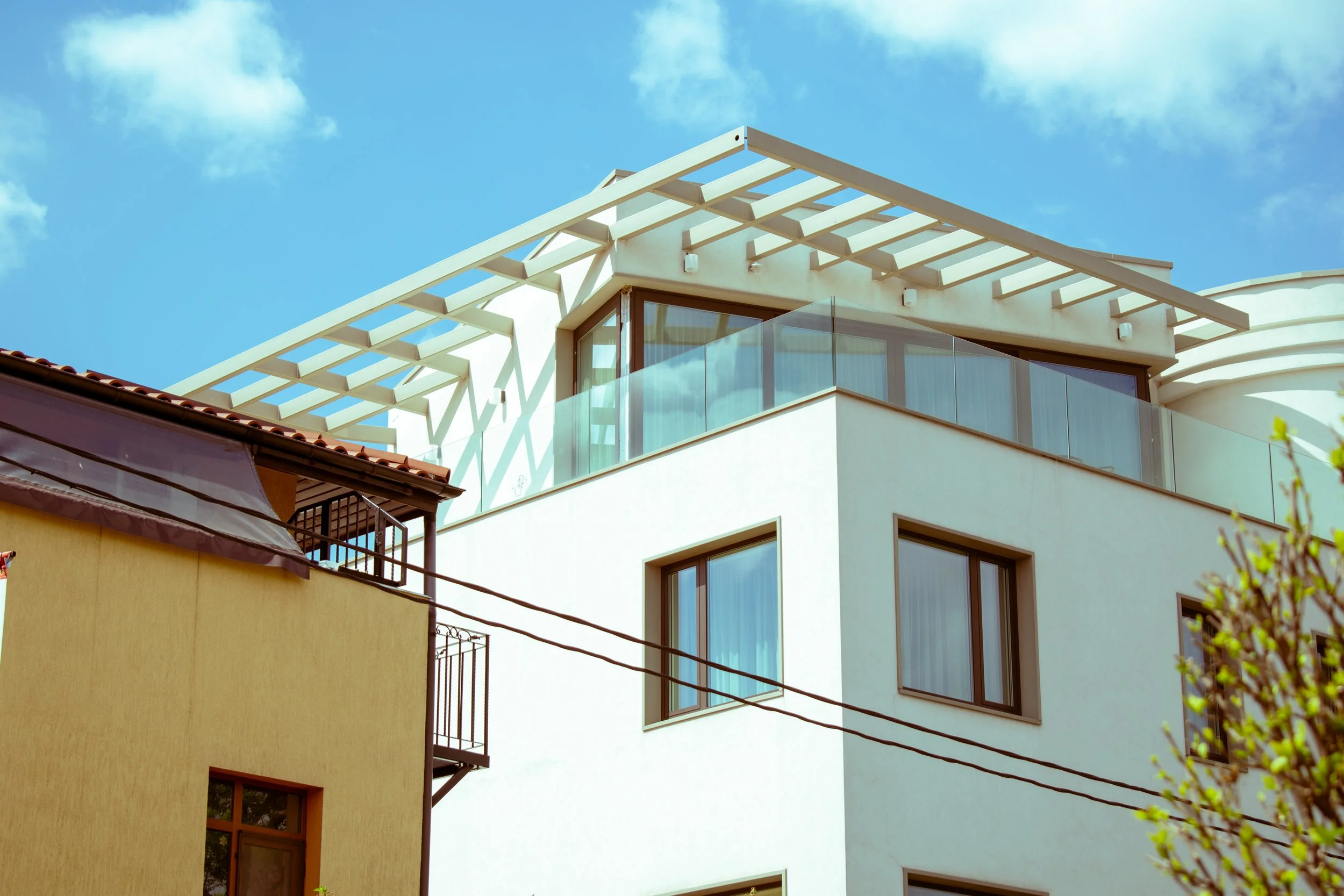 Modern residential building with a white facade, large windows, a glass balcony, and a rooftop pergola against a blue sky.