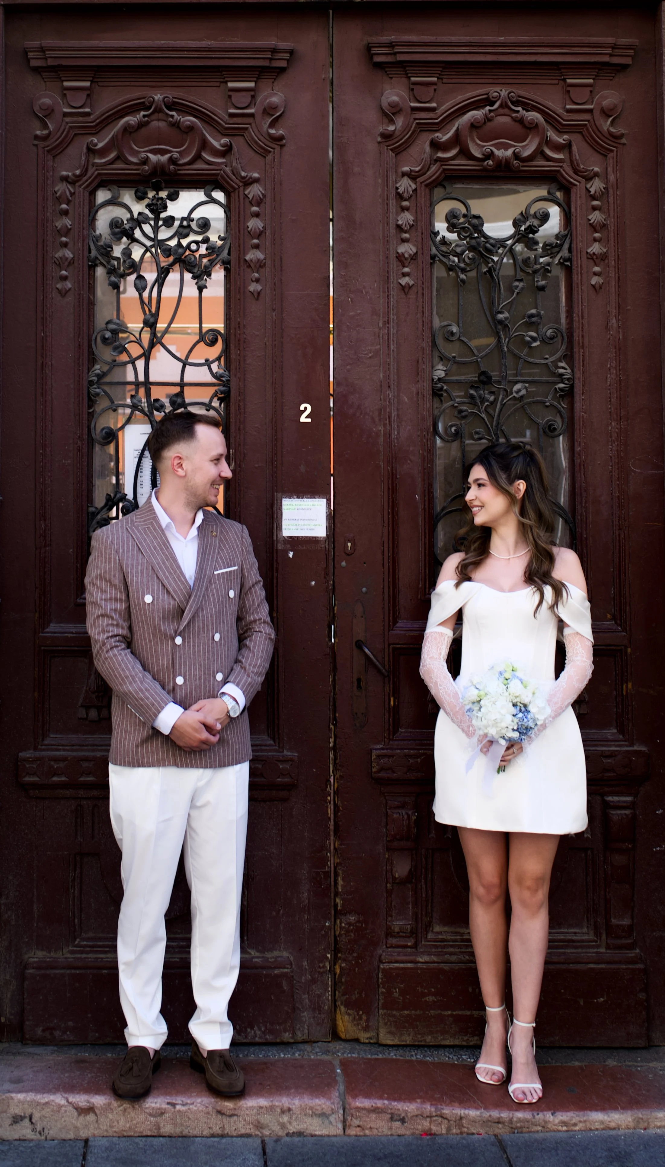 A man and woman dressed in wedding attire standing in front of ornate wooden doors, smiling at each other.