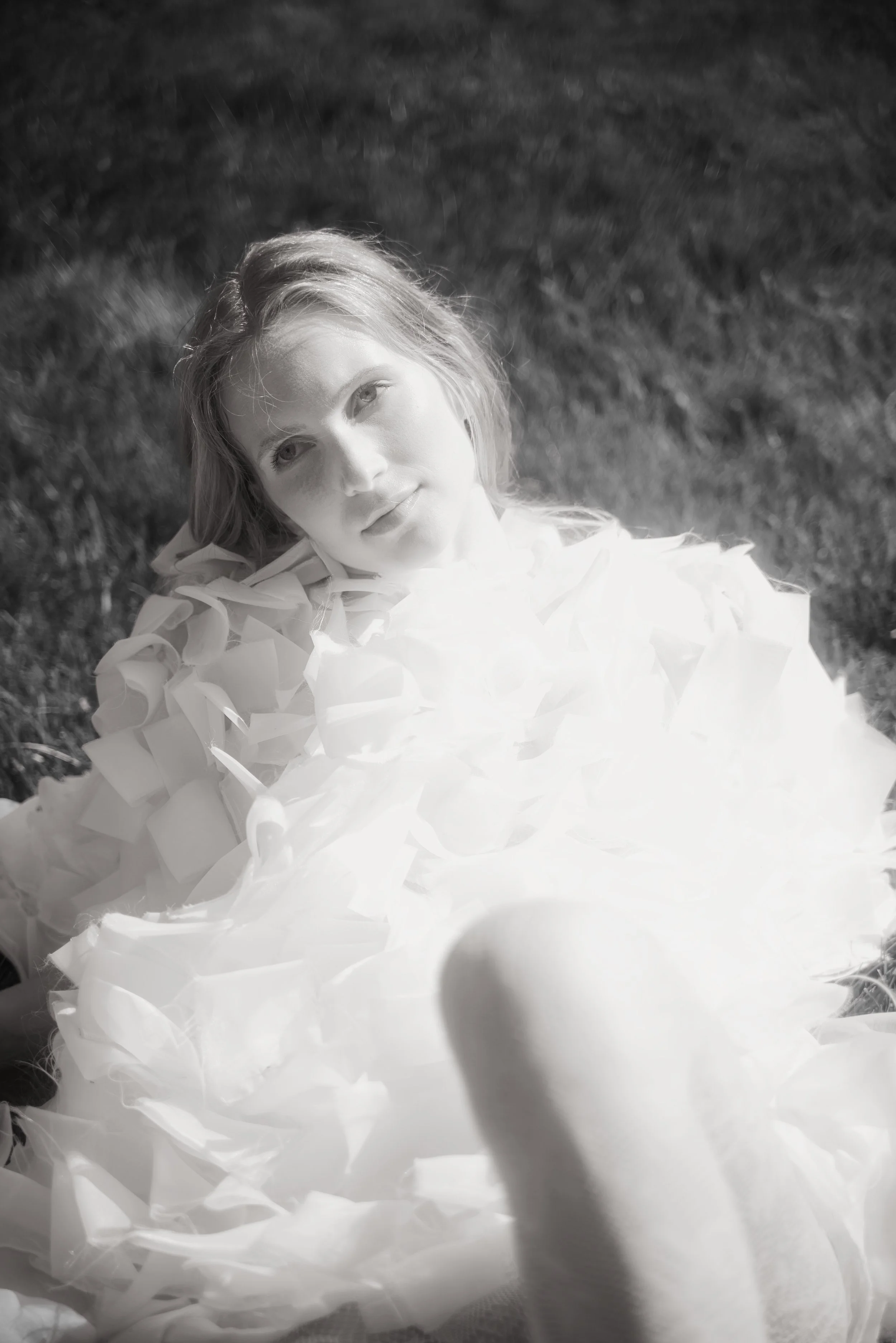 A young woman with light hair lies on grass, wearing a white dress with ruffled fabric, gazing thoughtfully at the camera in black and white.