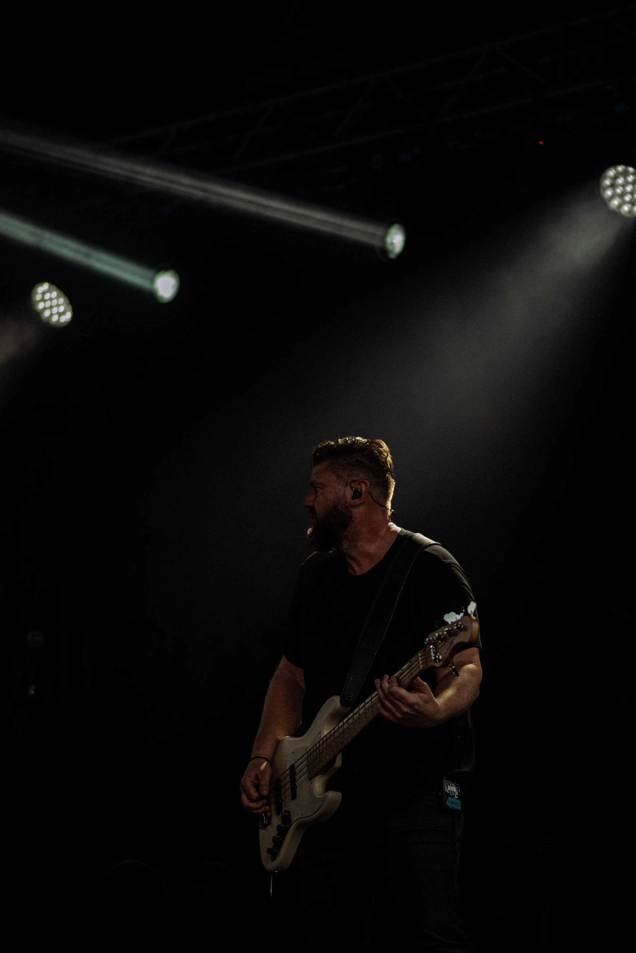 A man playing a bass guitar on stage in a dark concert setting with stage lights overhead.