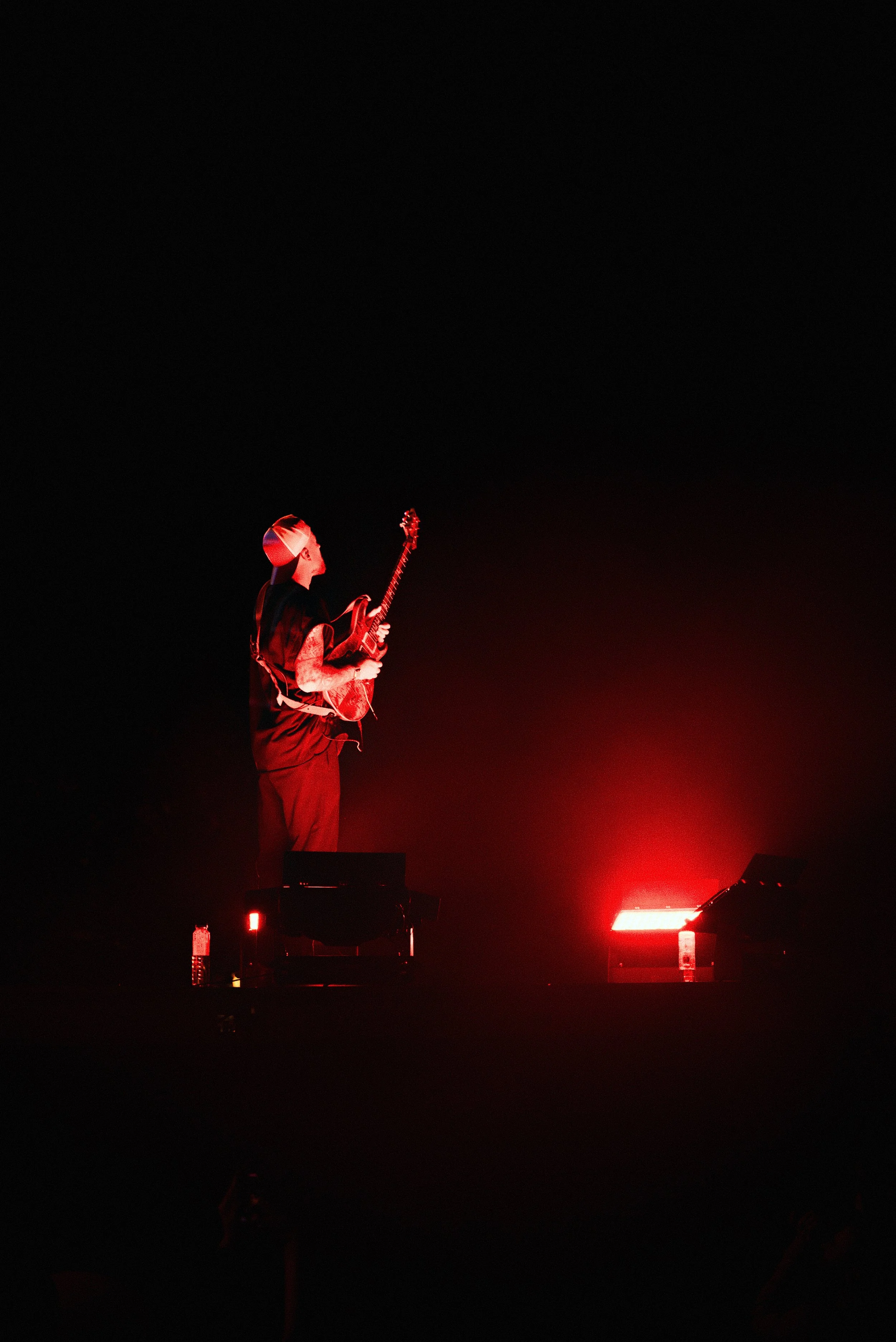 A musician playing an electric guitar on stage with red lighting, wearing a hat and sleeveless shirt.