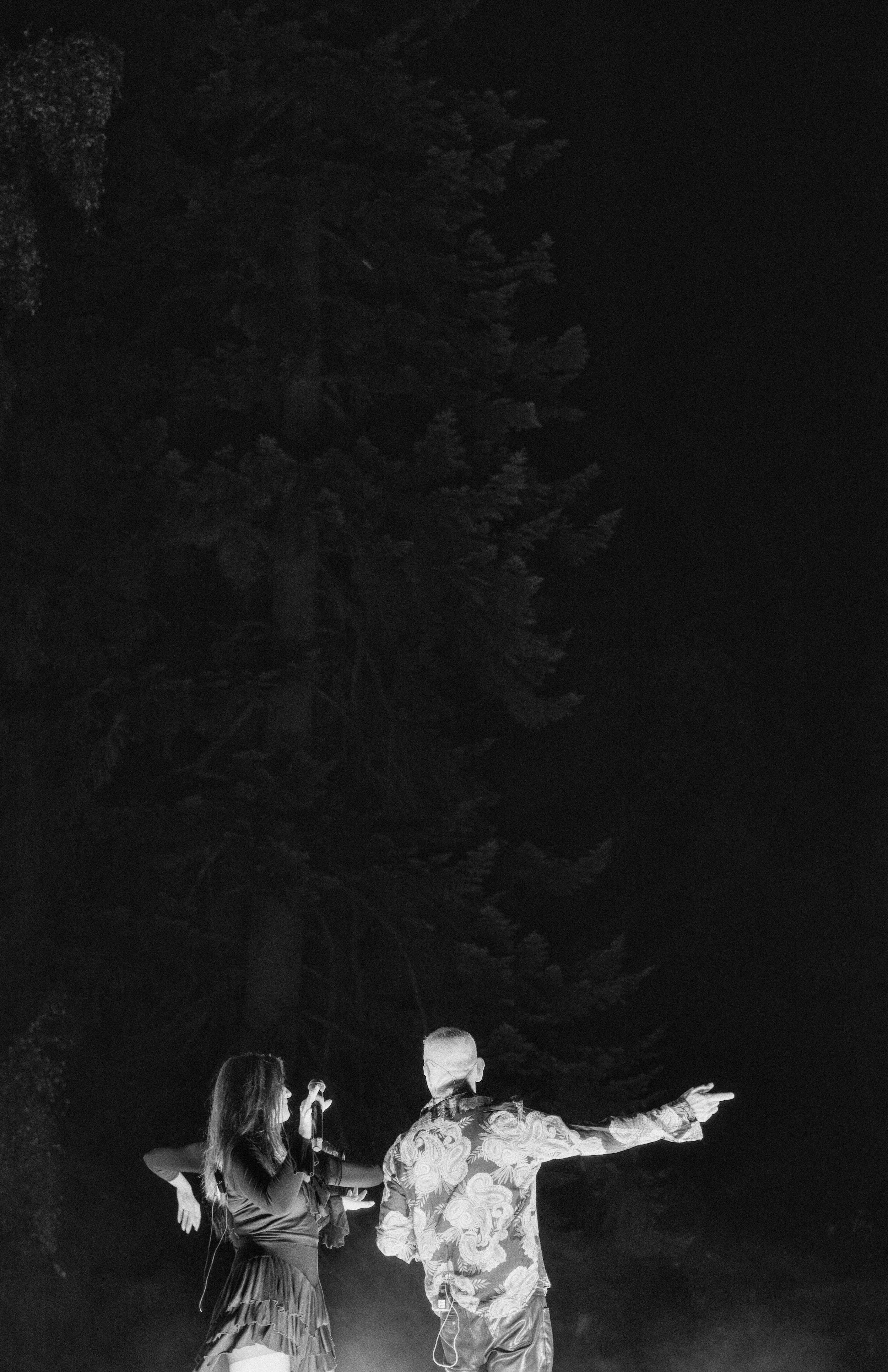 A man and a woman stand outdoors at night, with the man pointing into the distance. The woman is holding a microphone, and both are next to large trees in the dark.