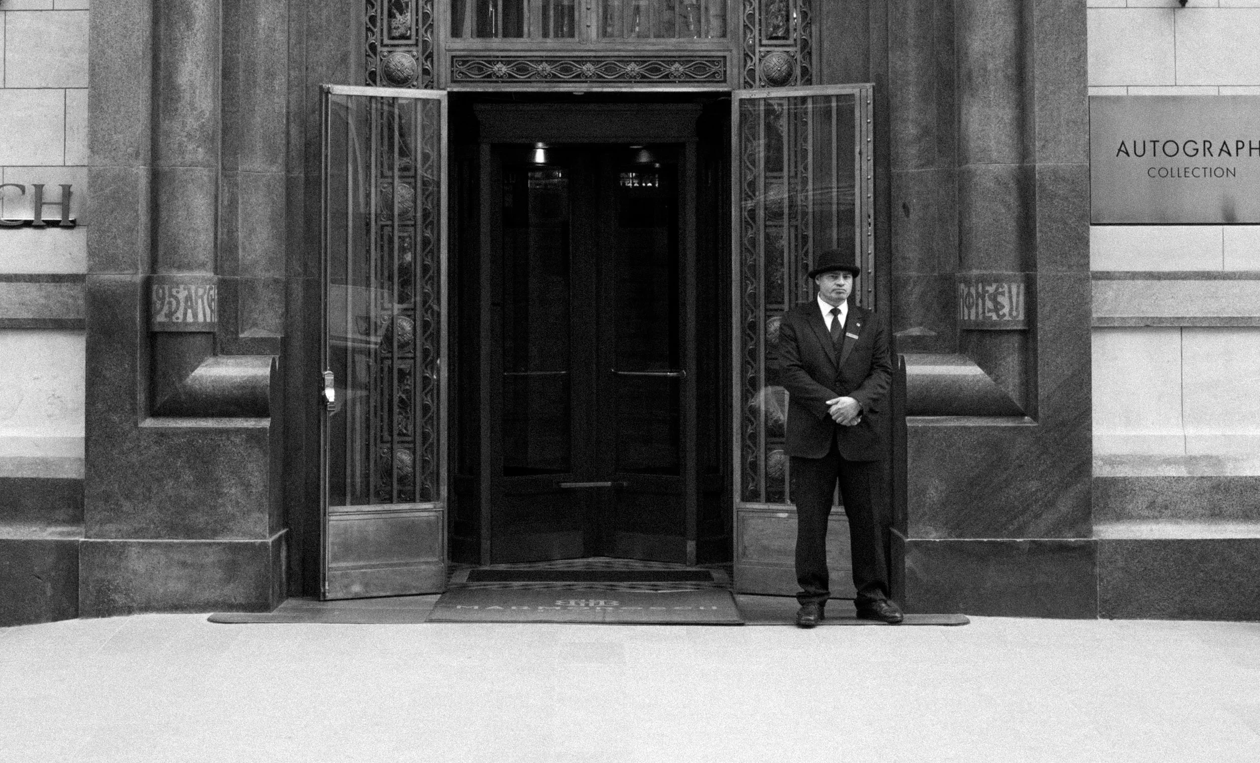 A doorman in a suit and hat standing outside the entrance of a building with ornate glass doors in black and white photo.
