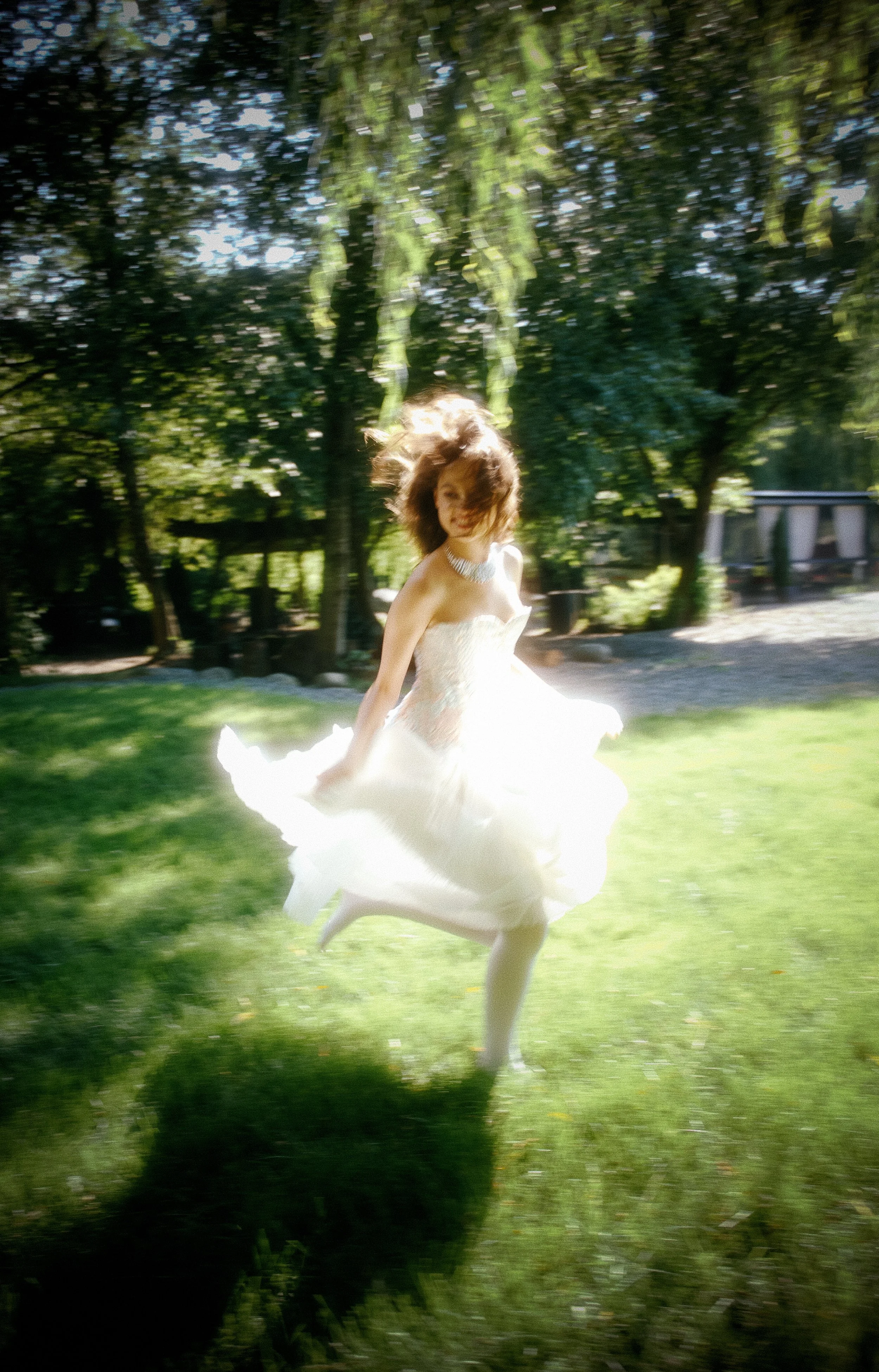 A woman in a white dress spinning outdoors on a grassy area surrounded by trees, with sunlight filtering through the leaves.