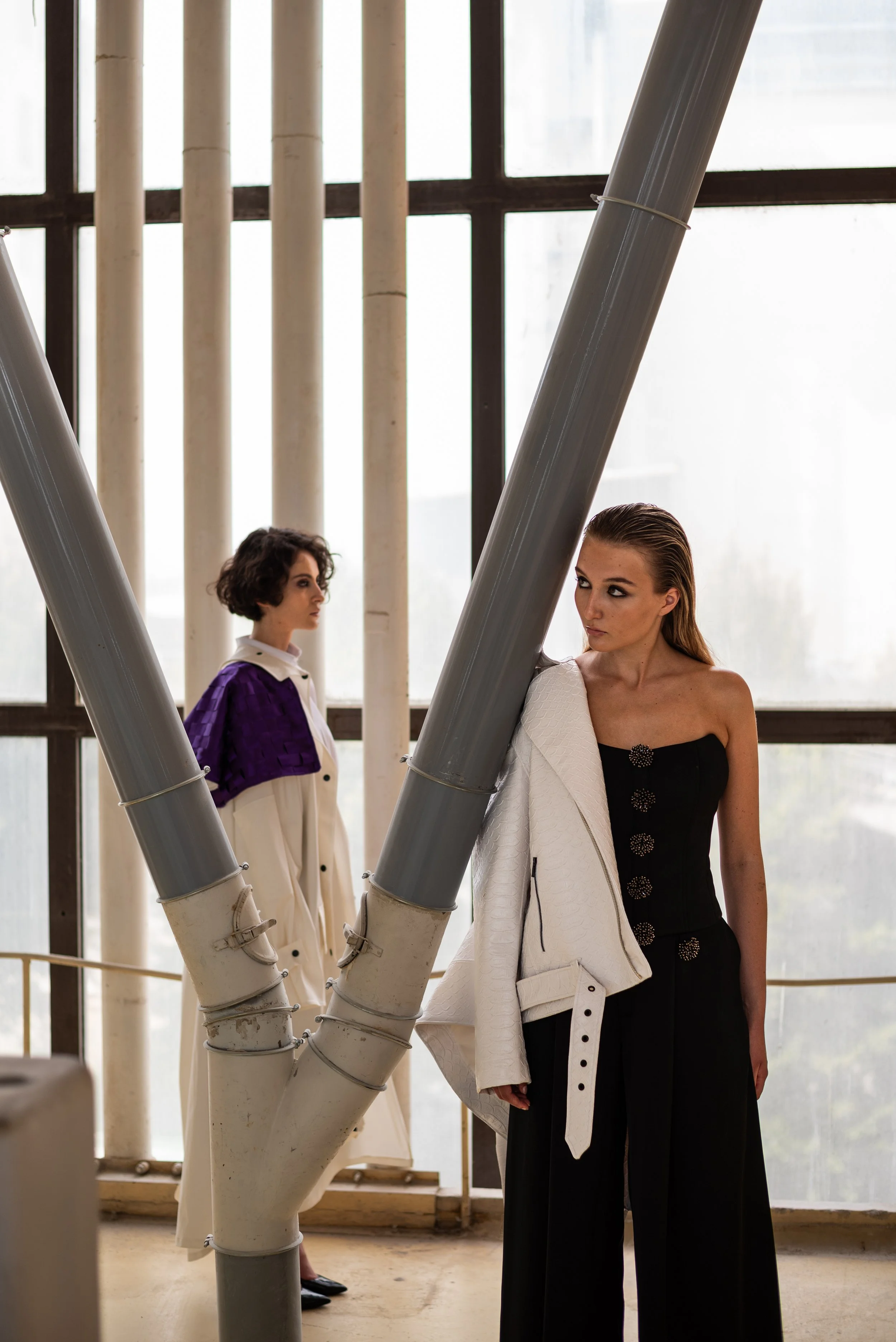 Two women dressed in fashionable clothing pose in front of large windows and industrial pipes in an indoor setting.
