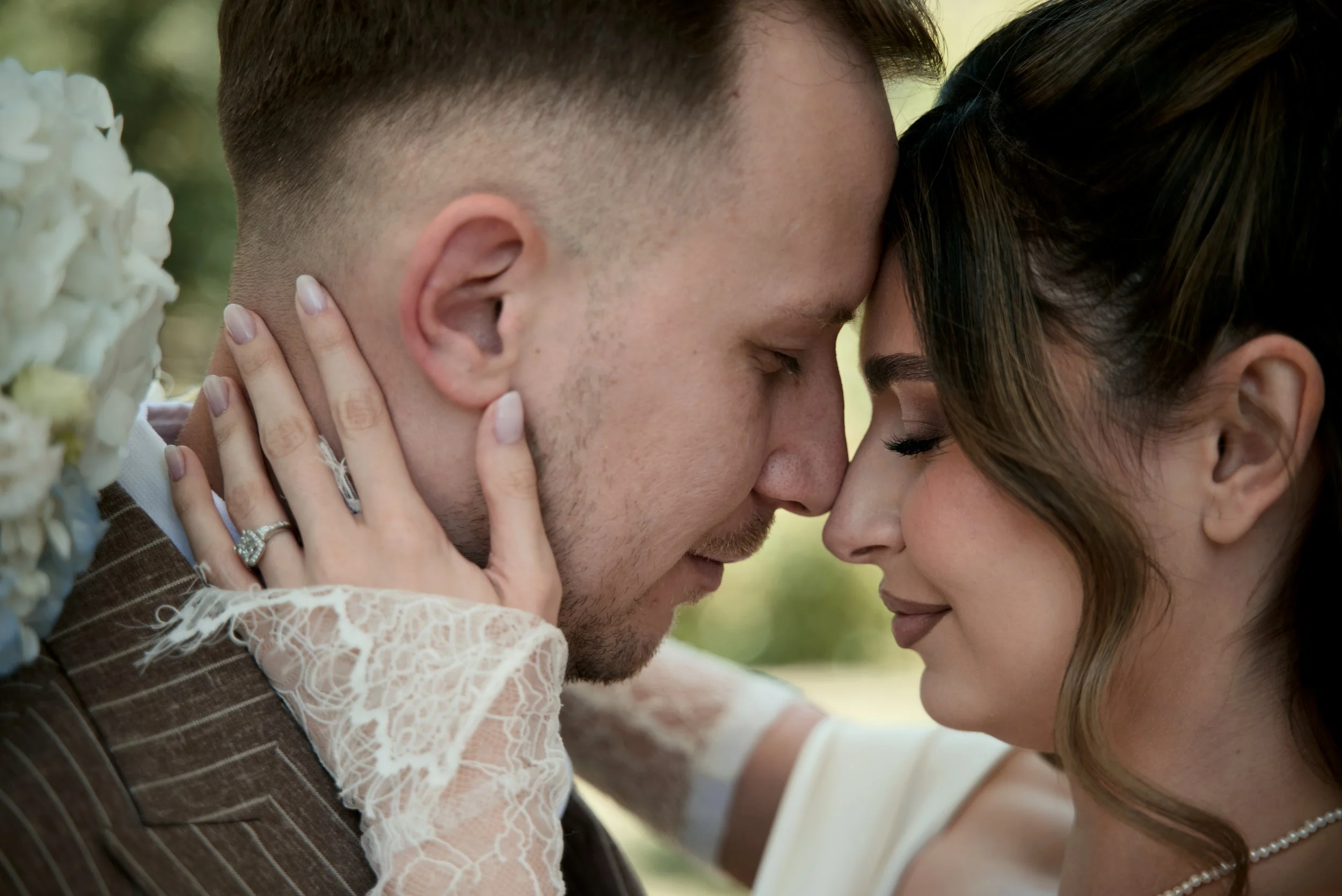 Close-up of a couple with their foreheads touching during a wedding ceremony, with the woman's hand visible resting on the man's face, showing her engagement ring and wedding band.